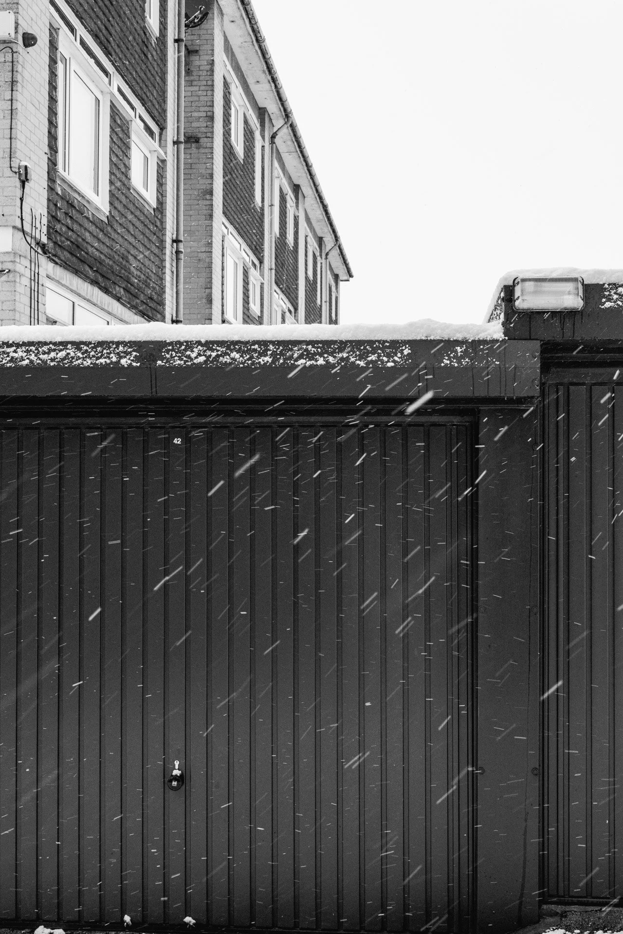 Black garage door with snow and falling snowflakes, residential building in the background, black and white photograph.