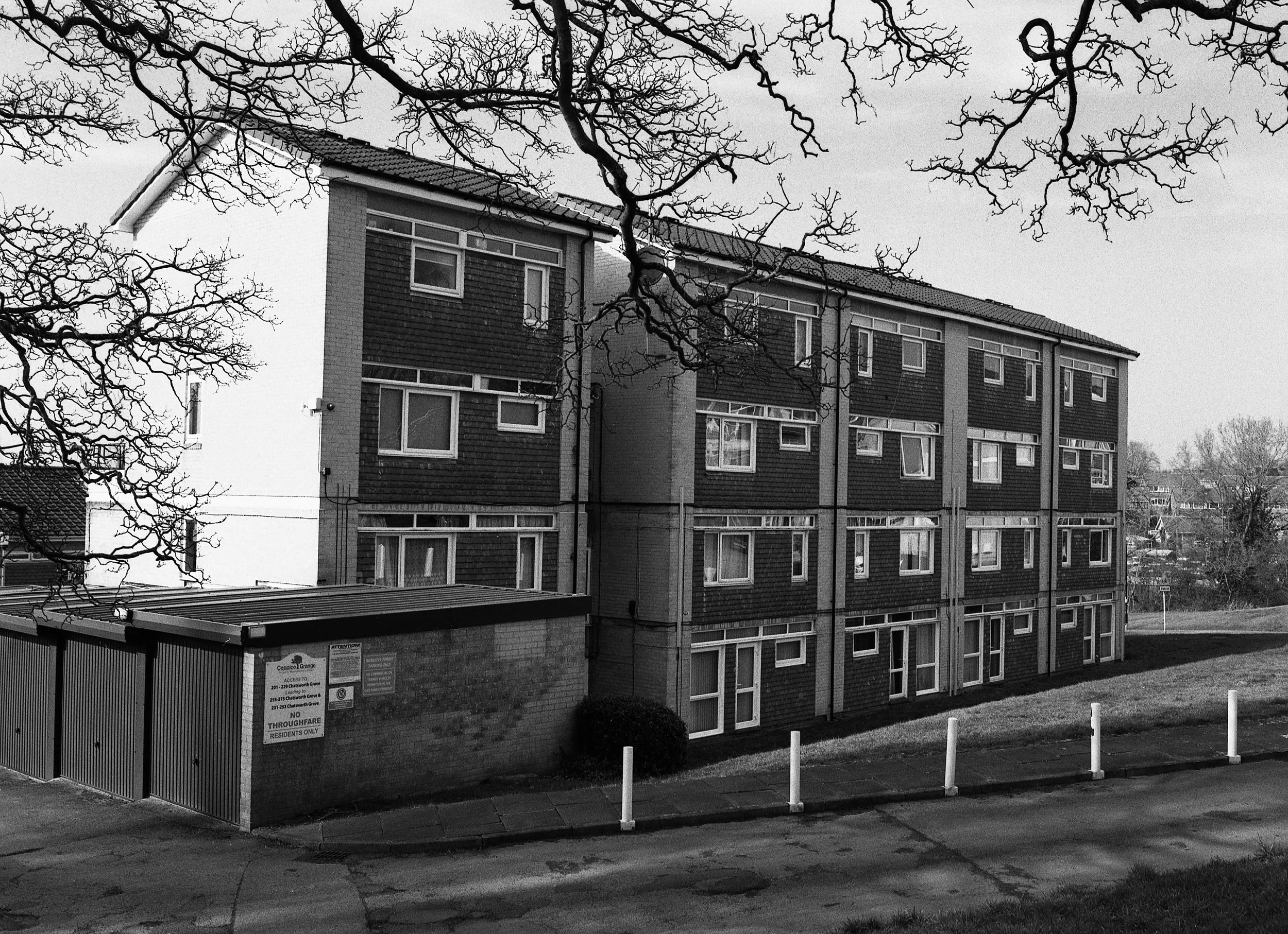 A black and white photo of a four-story apartment building with multiple windows, a pitched roof, and a plain brick exterior. In the foreground, leafless tree branches extend across the top of the image, and a small brick utility structure with a sig