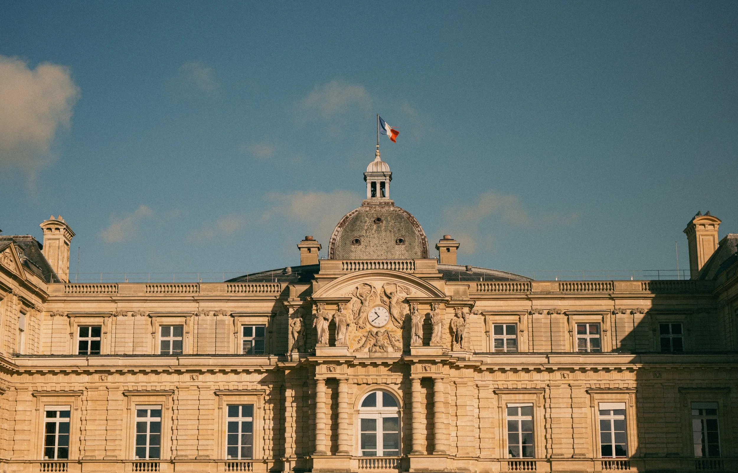 A historic European-style building with ornate architecture, topped with a clock and statues, and a French flag on the roof against a blue sky with some clouds.