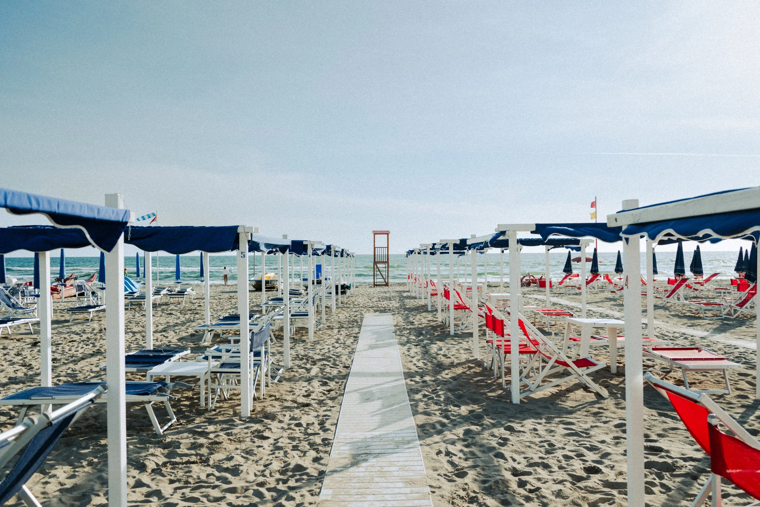 Empty beach with blue and red striped lounge chairs and blue umbrellas, a wooden walkway leading to the ocean, and a lifeguard tower in the background.
