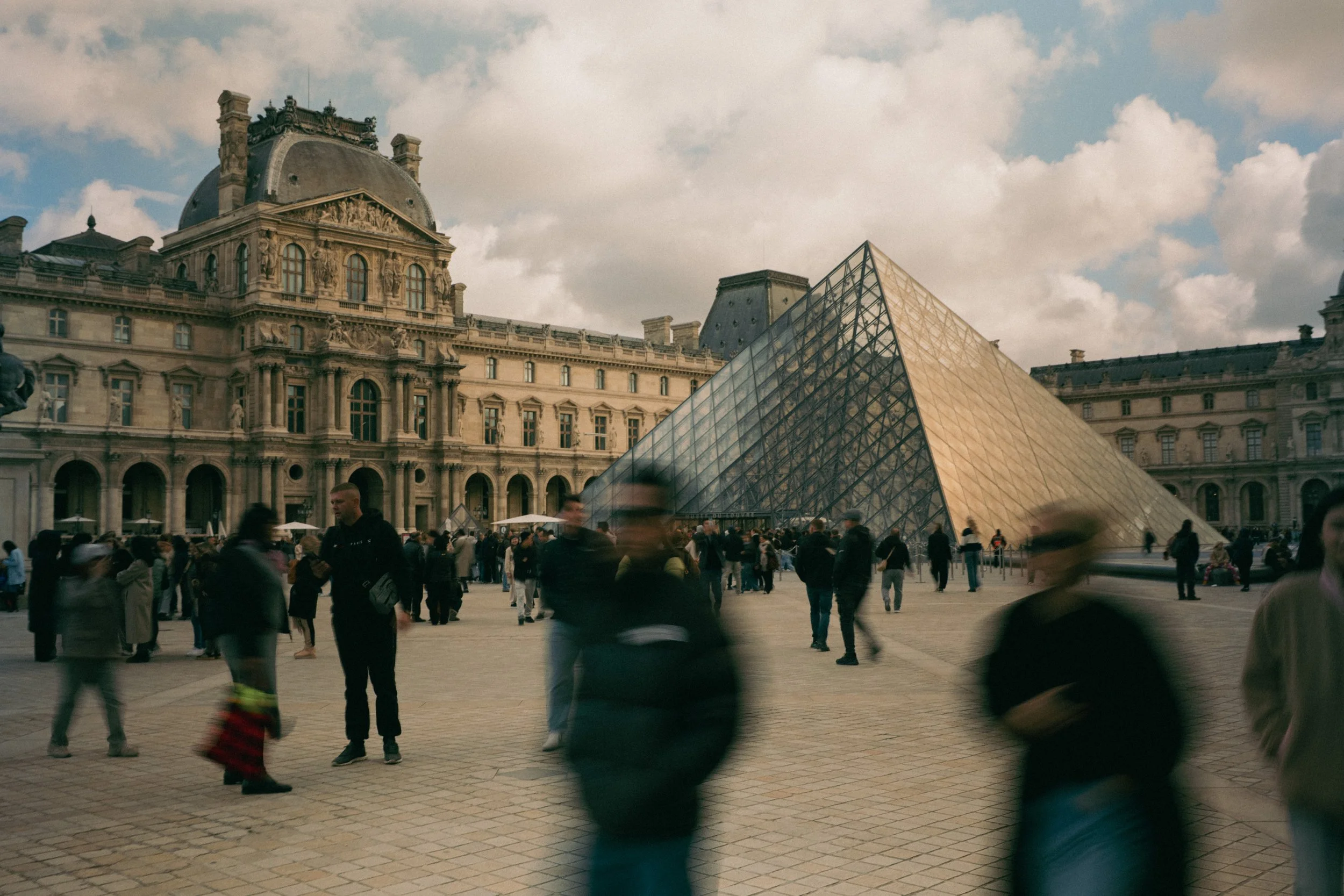 People walking around the outdoor Louvre Museum courtyard with the glass pyramid and historic building facades in Paris, France.