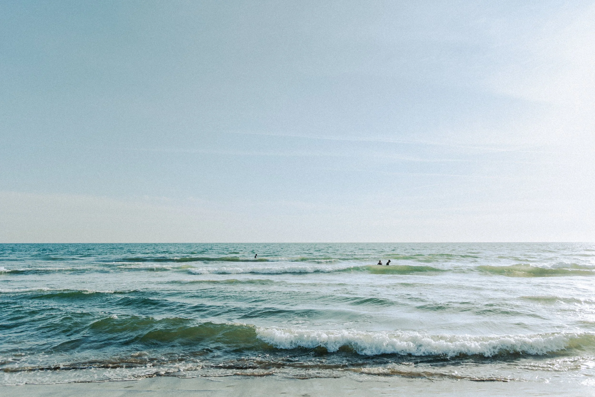 A calm ocean shoreline with gentle waves and a clear sky, two people are in the water near the horizon.