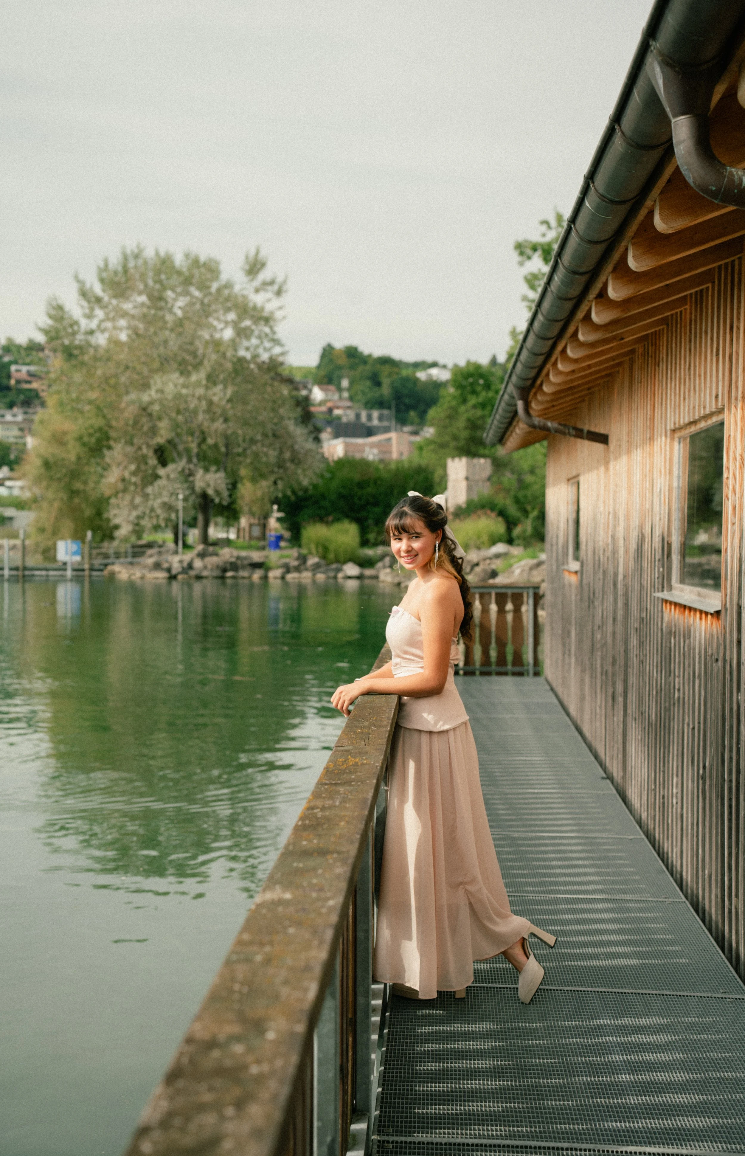 A young woman in a strapless dress and high heels standing on a metal walkway by a wooden building, smiling and looking at the camera, with a pond and trees in the background.