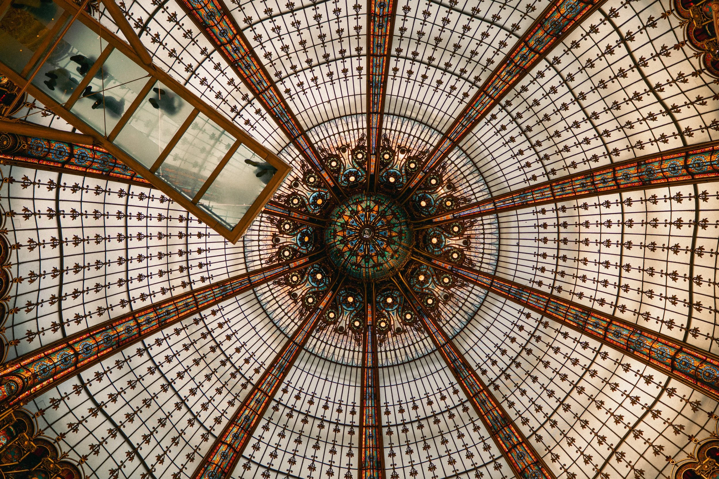 Looking up at the ornate glass dome ceiling with intricate stained glass patterns and metal framework, featuring a rectangular skylight with reflection of indoor lights.