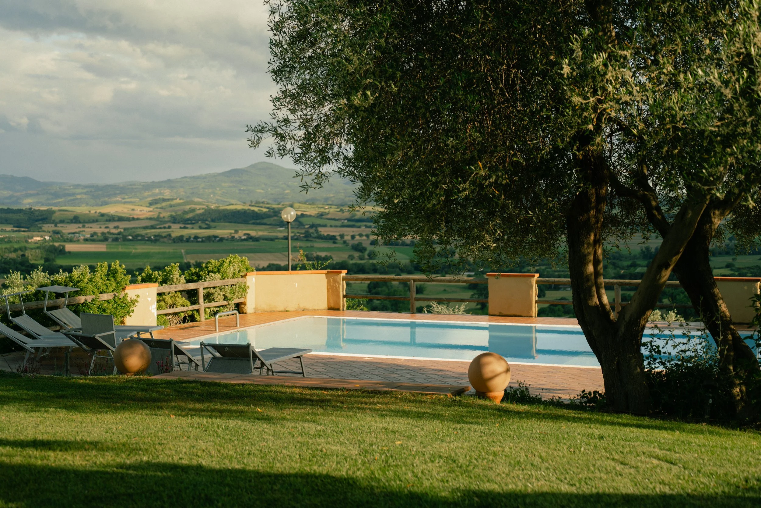 view of a swimming pool in a lush outdoor setting with lounge chairs, large ceramic pots, and a sprawling landscape of green fields and hills in the background, under a partly cloudy sky.