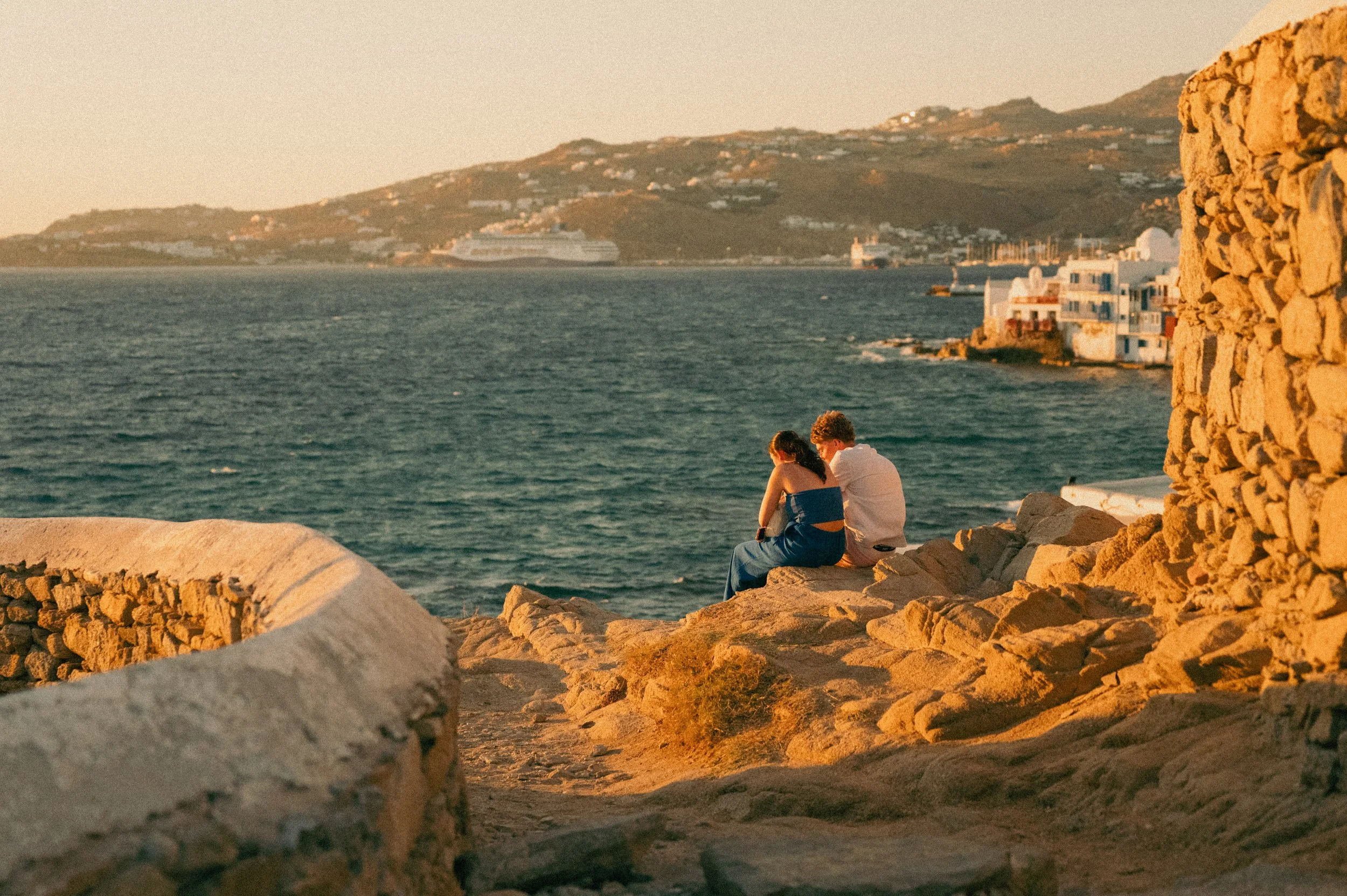 A couple sitting on rocks by the sea during sunset, with distant hillside buildings and a cruise ship.