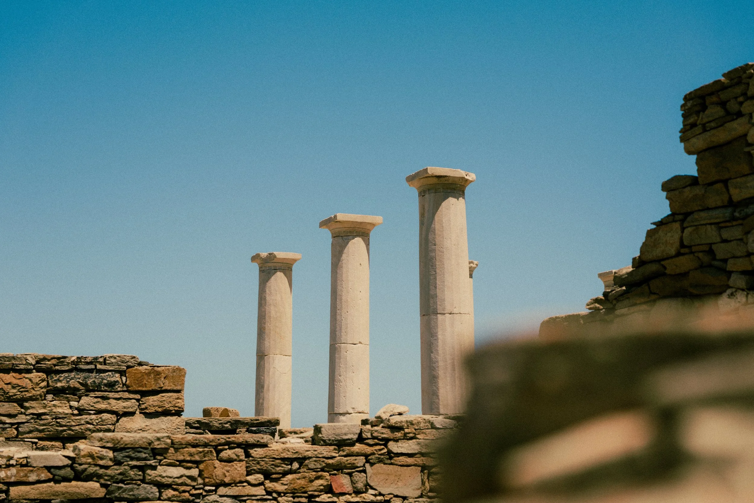 Ancient stone ruins with four tall columns and a partial stone wall against a clear blue sky.