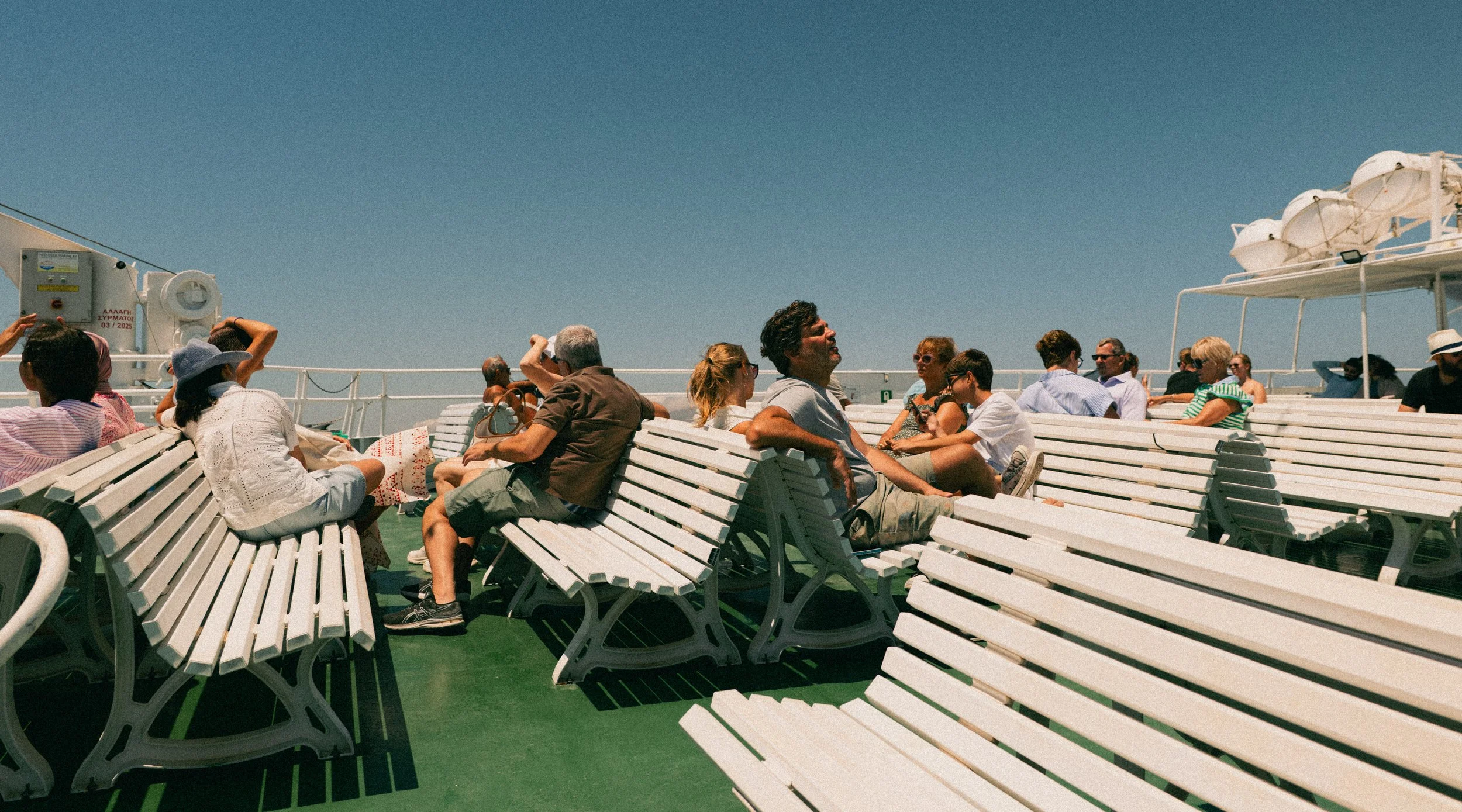 People relaxing and sitting on white benches on a ferry boat under a clear blue sky.