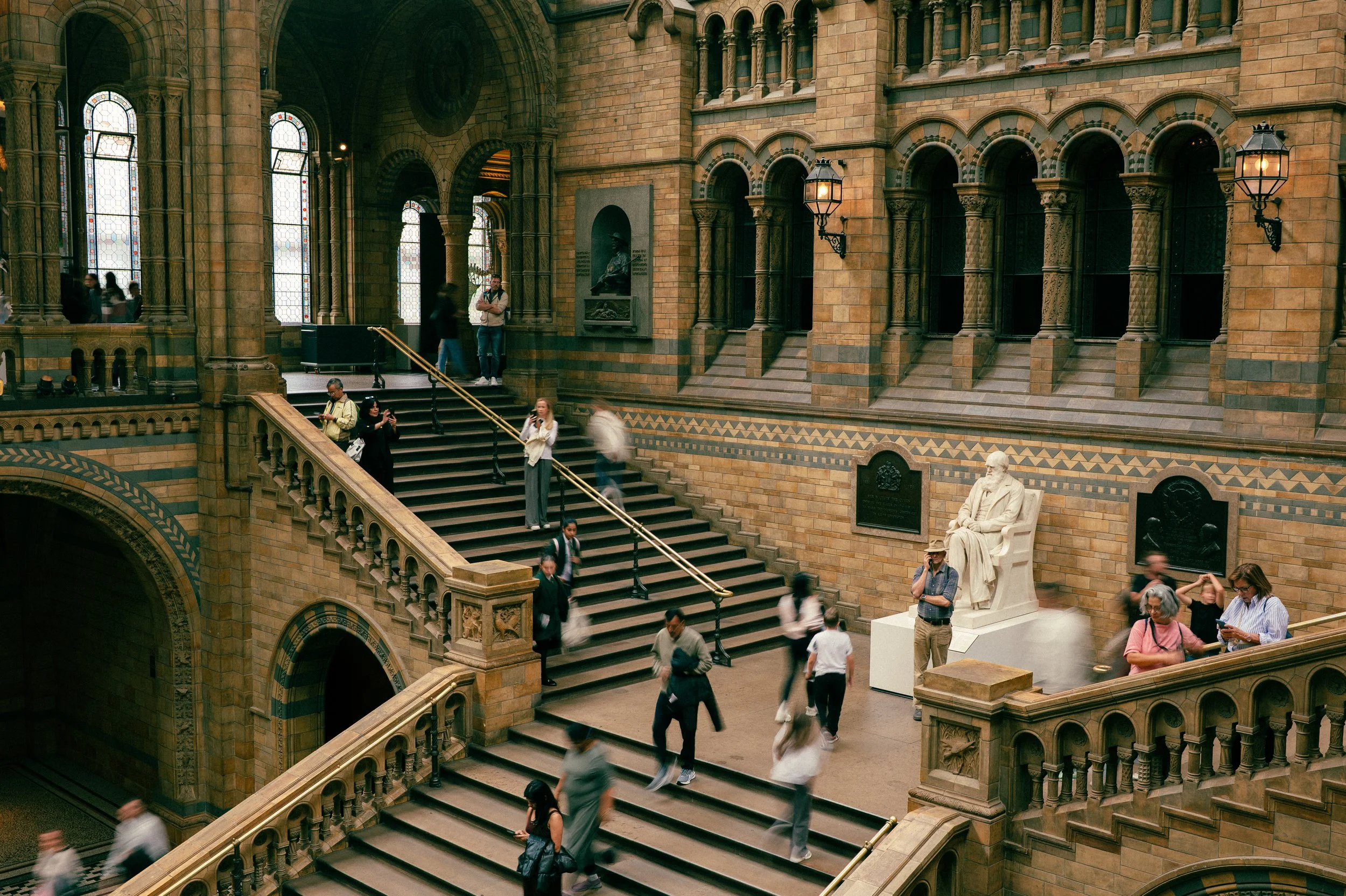 Interior view of a historic building with grand staircase, arched windows, stone walls, and a statue of a seated man.