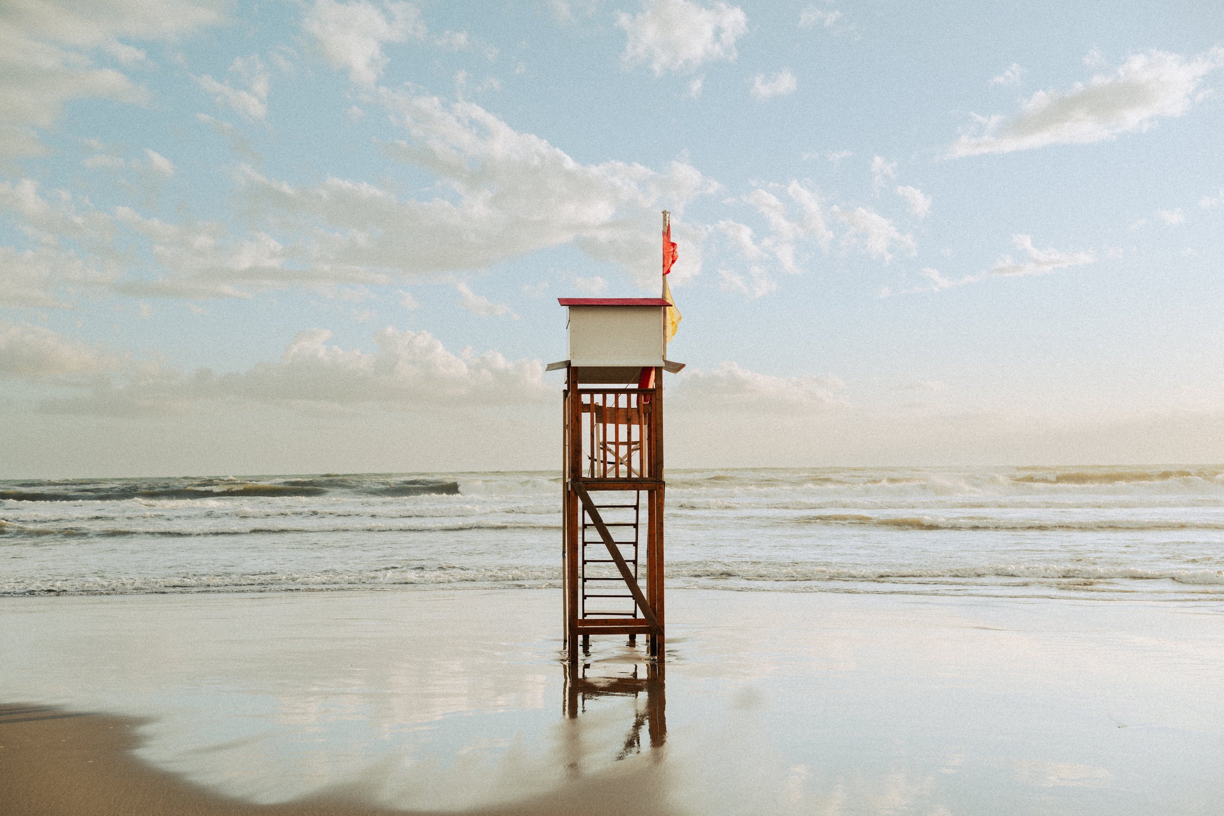A lifeguard tower on a sandy beach with ocean waves and a partly cloudy sky in the background.