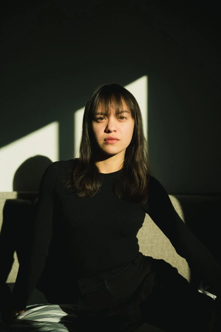 A young woman with medium-length brown hair and bangs, wearing a black long-sleeve shirt and black pants, sitting on a gray couch in a room with dark walls. She is looking directly at the camera with a neutral expression. Sunlight streams in through a window, creating geometric shadows on the wall behind her.