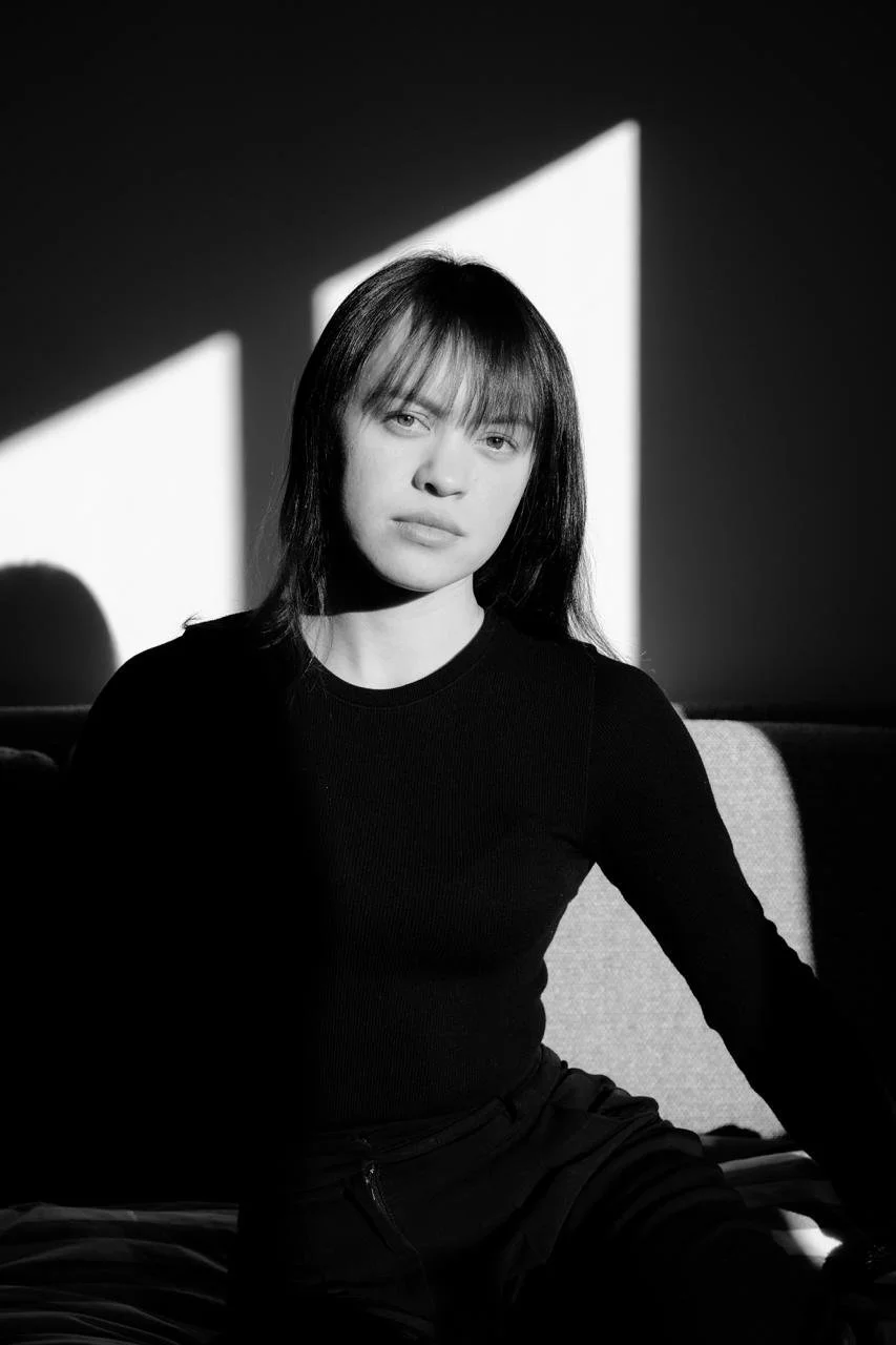 Black and white photo of a woman with shoulder-length hair and bangs, sitting in front of a wall with geometric light patterns.