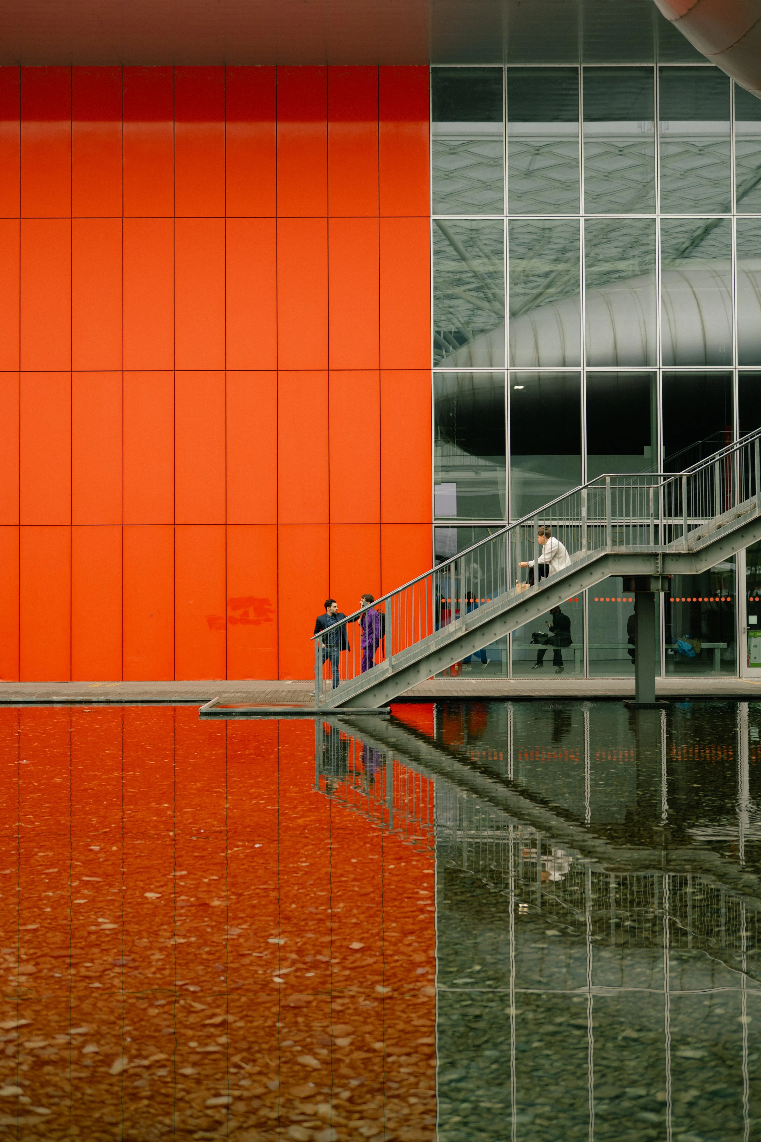 People walking on a glass staircase outside a modern building with orange and glass walls, reflected in a water feature below.