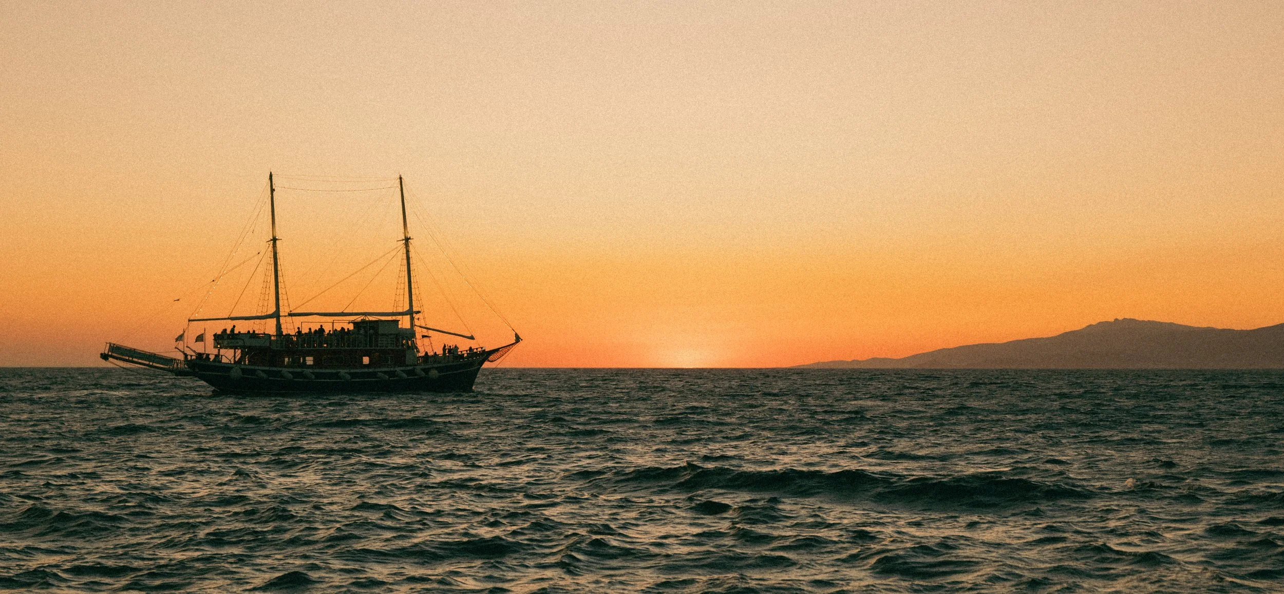A boat sailing on the ocean during sunset with a mountainous landmass in the distance.