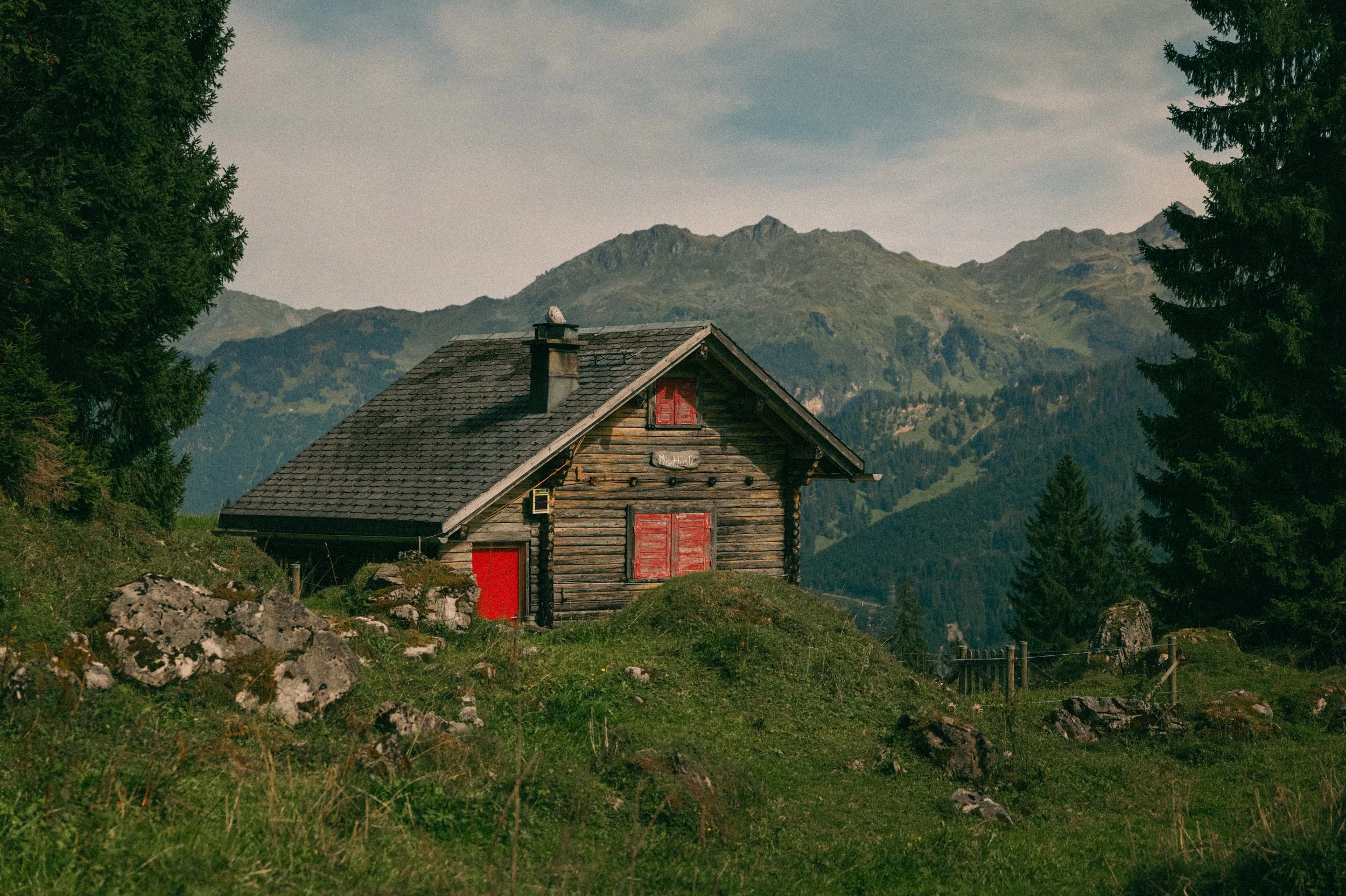 A rustic mountain cabin with red shutters and door, surrounded by green grass and rocks, with pine trees and mountains in the background under a partly cloudy sky.