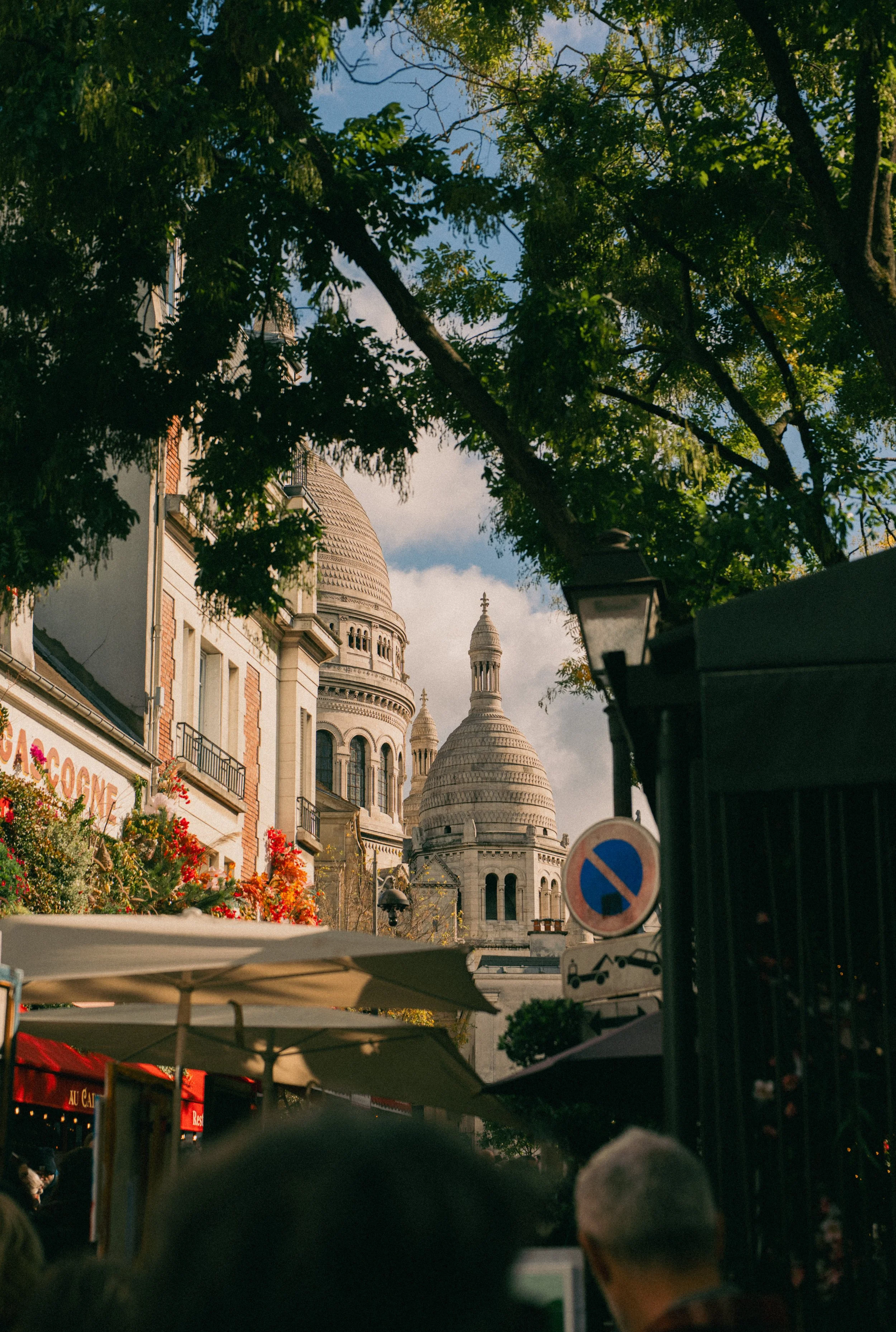 Parisian street scene with outdoor cafes and the Sacré-Cœur Basilica in the background, trees overhead, and a blue sky with clouds.