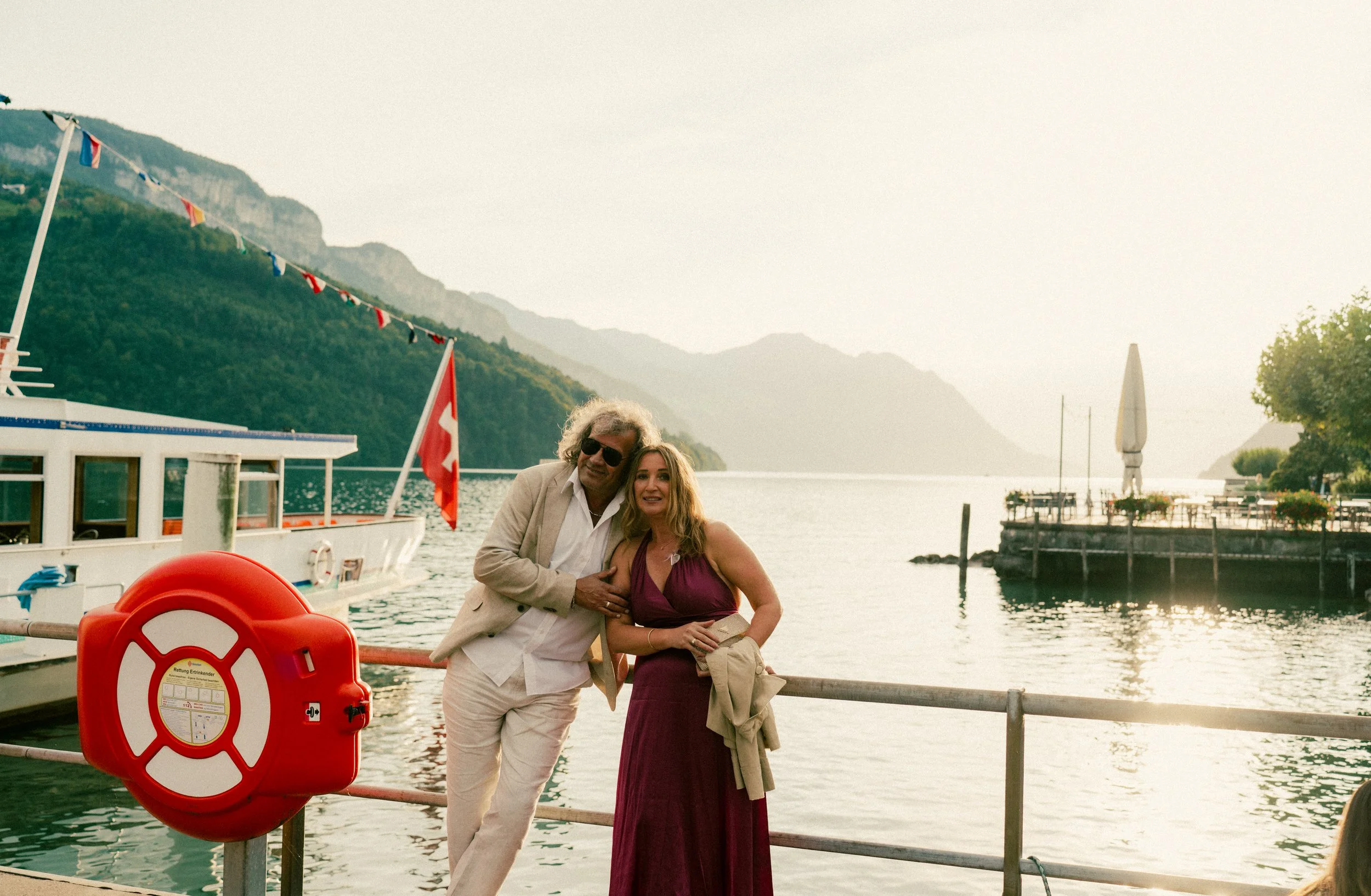 A man and woman posing on a dock by the water with boats, mountains, and a scenic lake in the background during sunset.