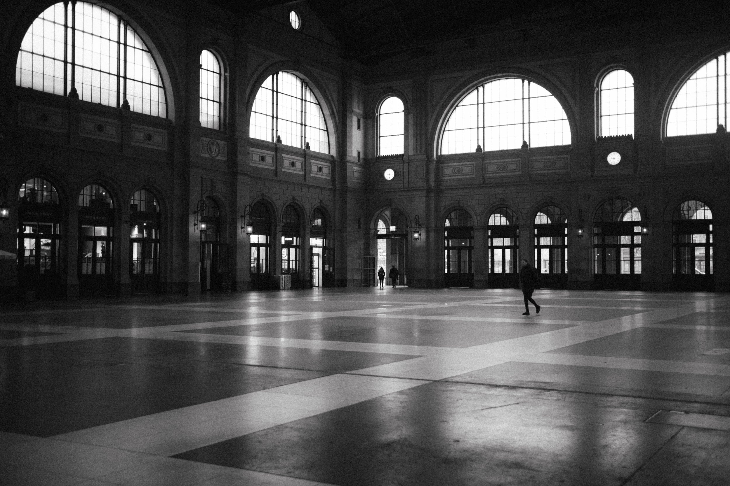 Interior of a historic train station with large arched windows, sparse lighting, and a spacious open floor with a few people walking.