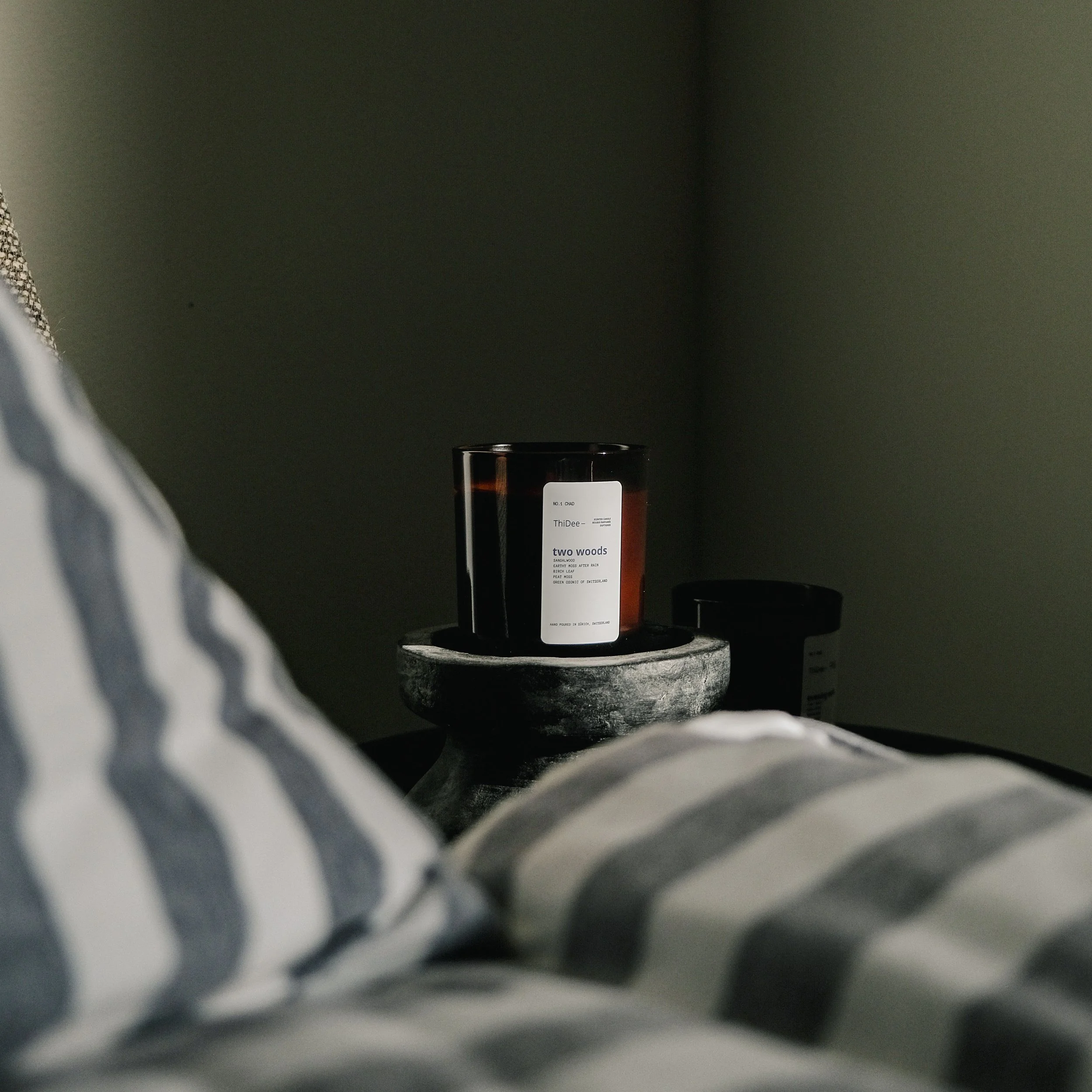 A candle labeled 'two woods' on a bedside table, partially visible in a bedroom with striped bedding and a plain wall background.