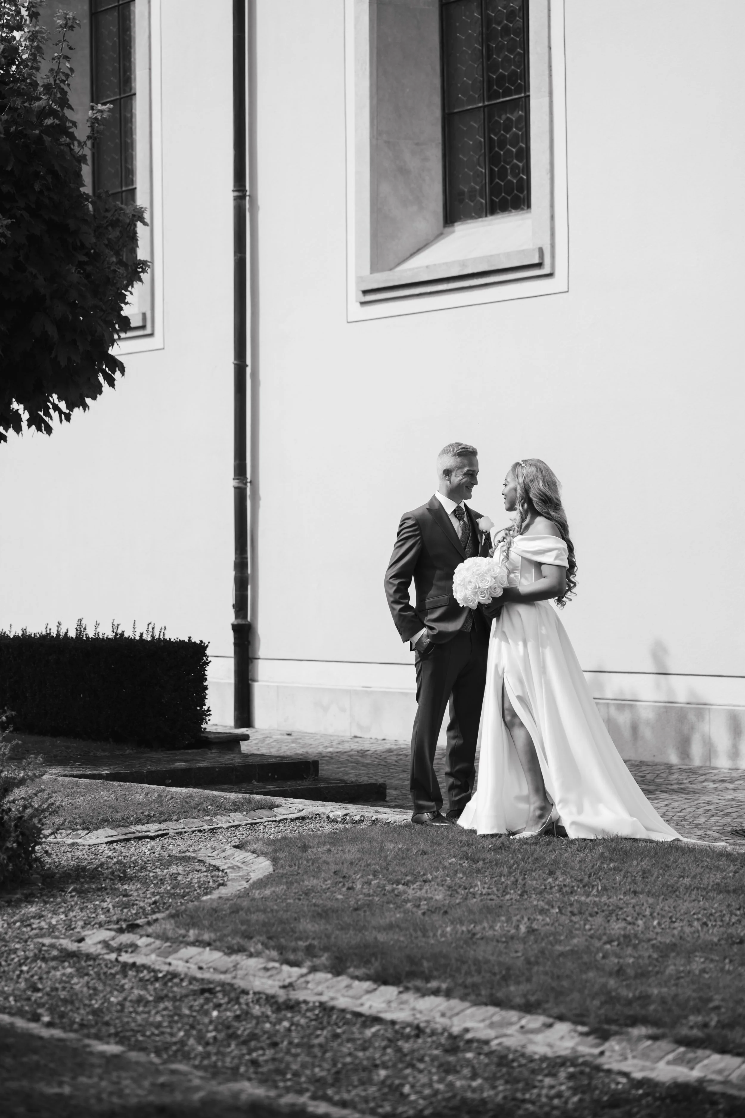 A black-and-white photo of a bride and groom standing outdoors near a building, smiling and looking at each other. The bride is holding a bouquet of flowers, and the groom is dressed in a suit.