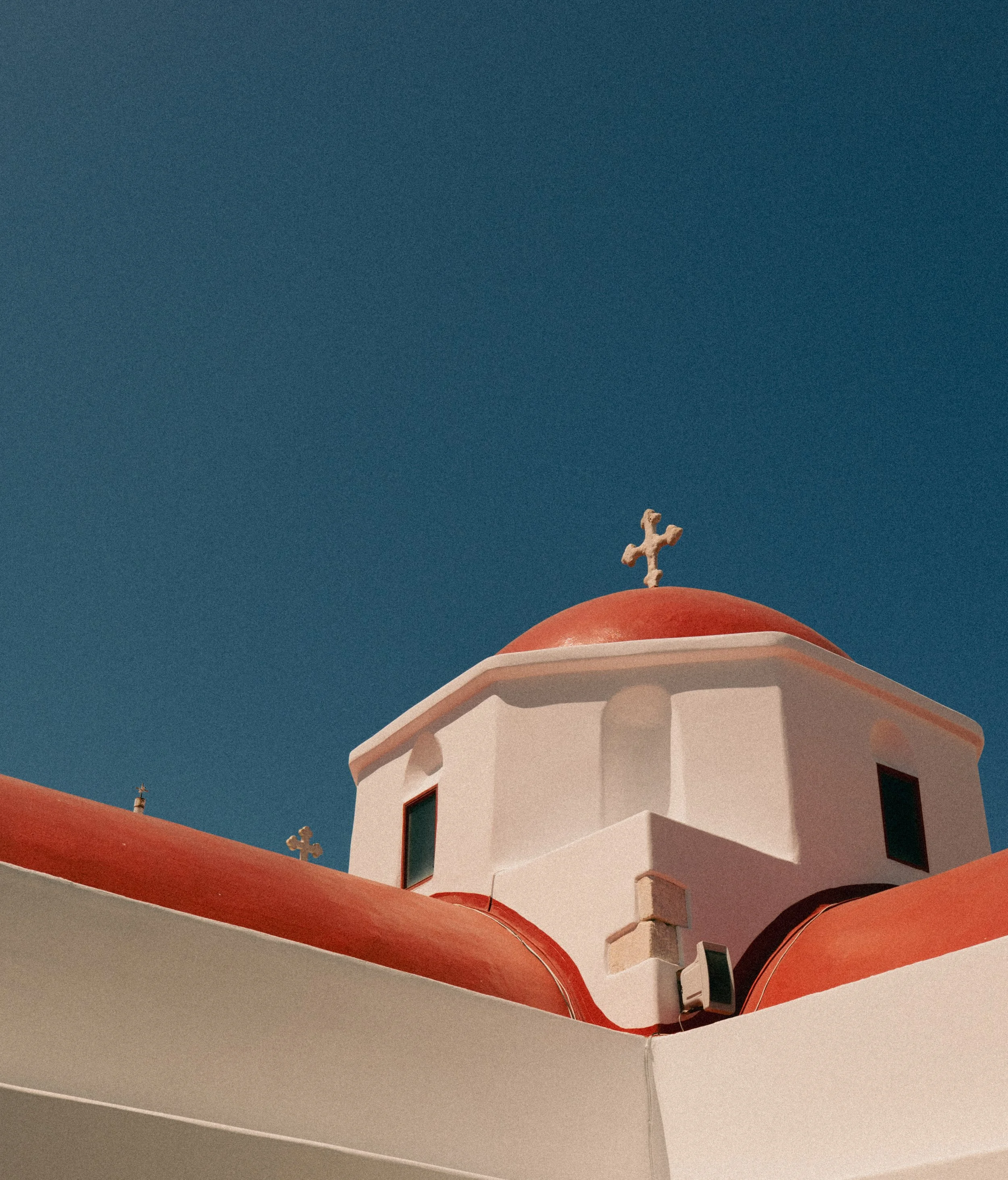 A white church building with red domed roofs and a cross on top, set against a clear blue sky.