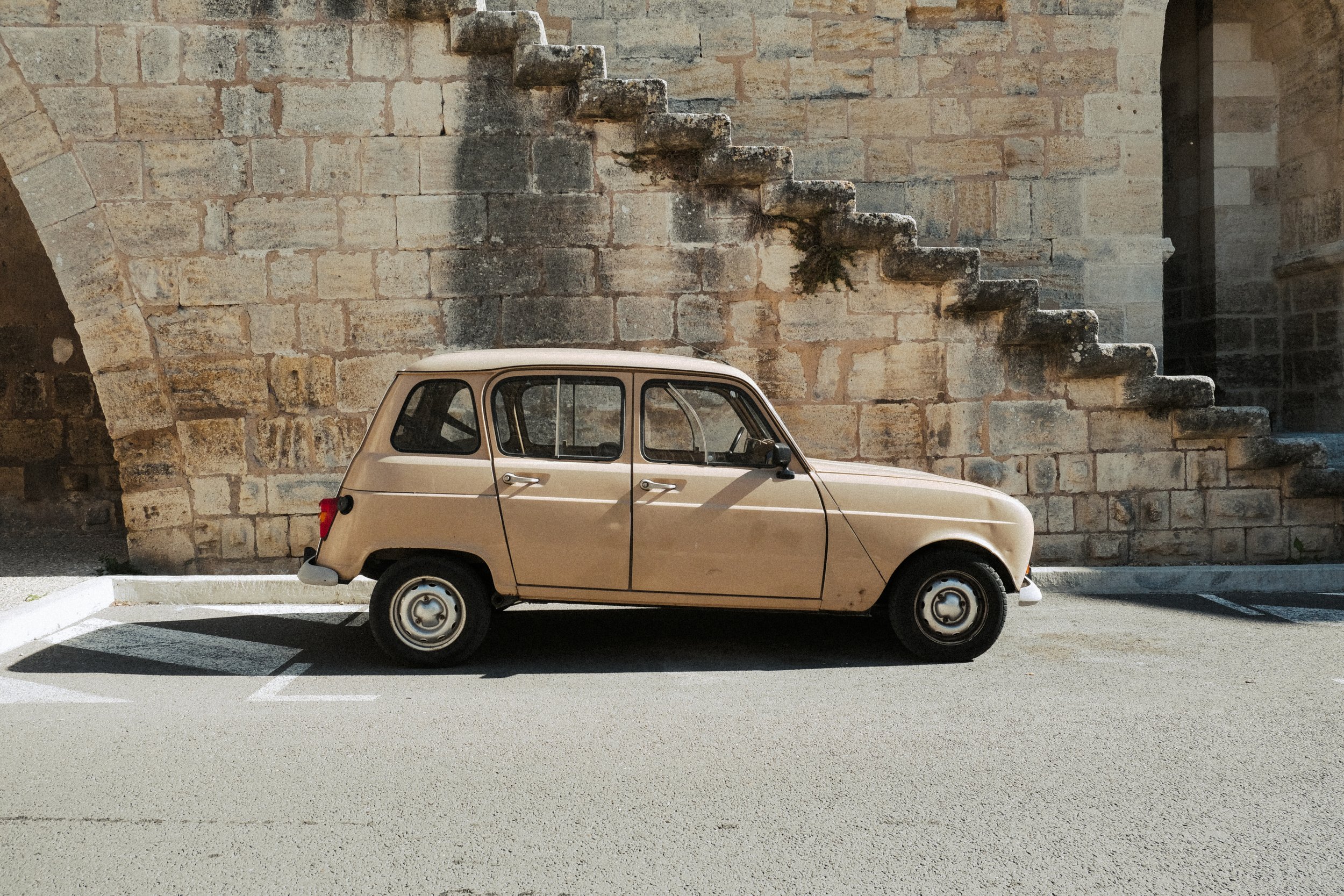 A vintage beige car parked in front of an old stone wall with stone stairs on the right side.