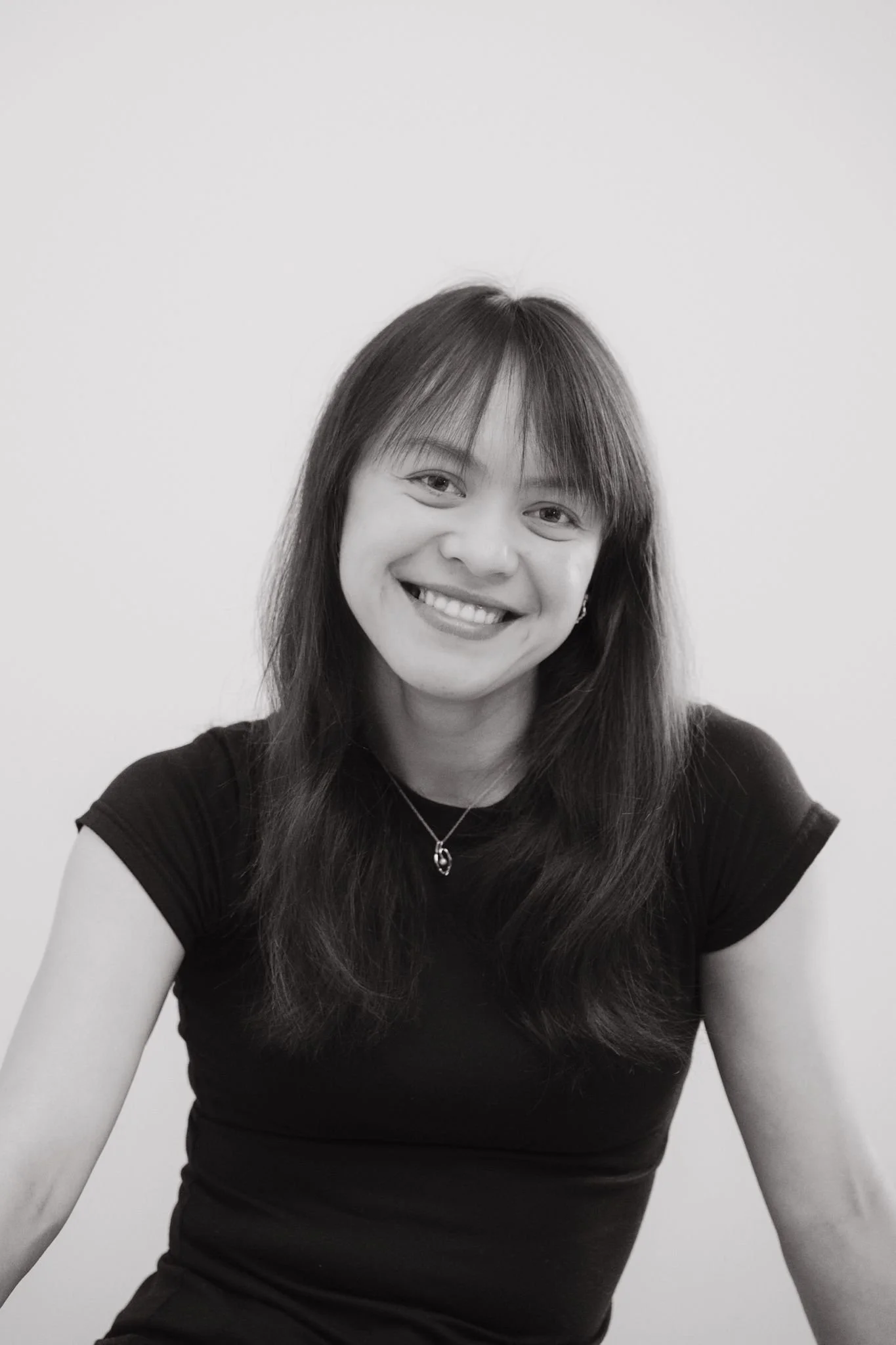 A smiling woman with straight shoulder-length hair and a necklace, wearing a black t-shirt, looking at the camera against a plain background.