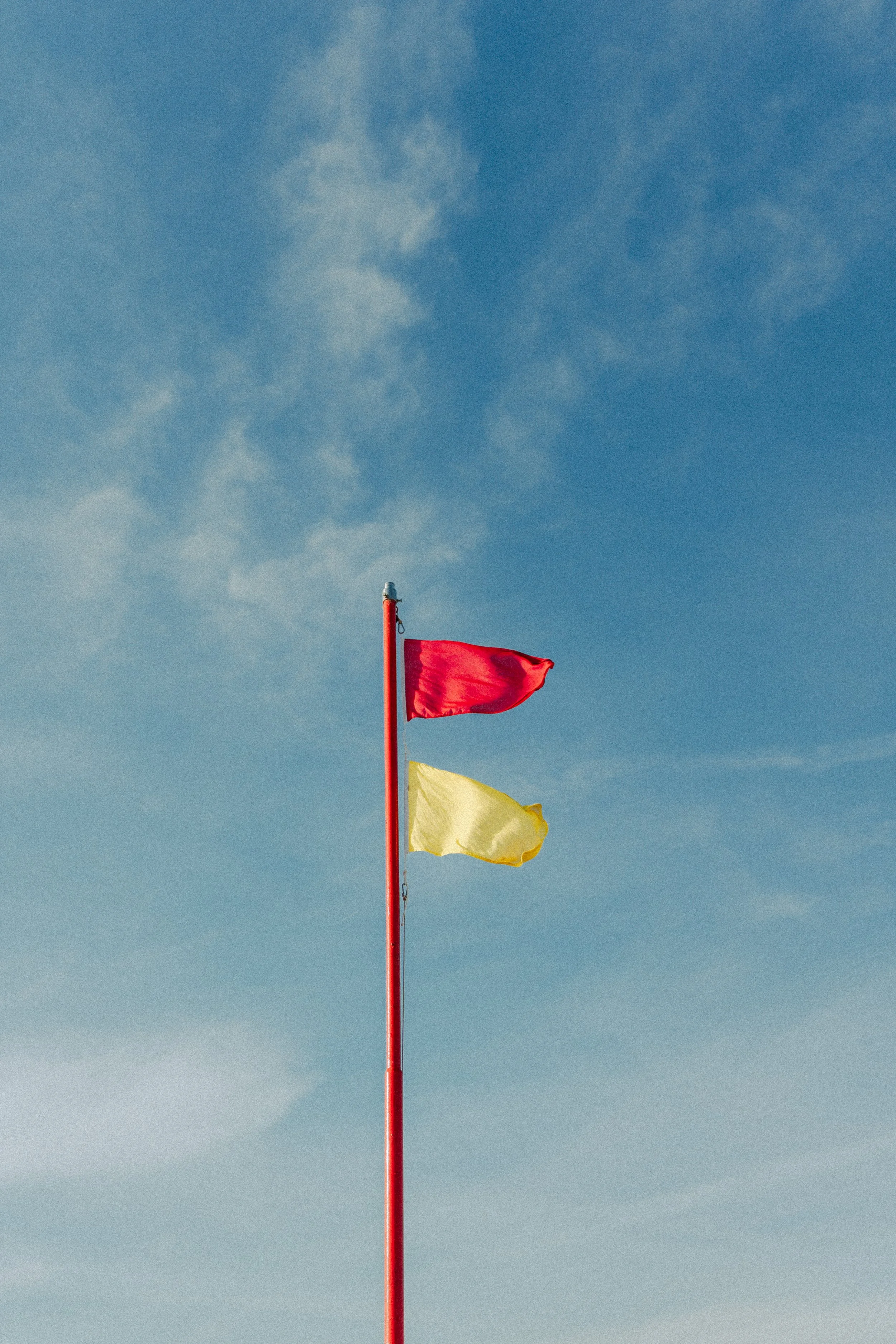 A red flag and a yellow flag on a red pole against a blue sky with some clouds.