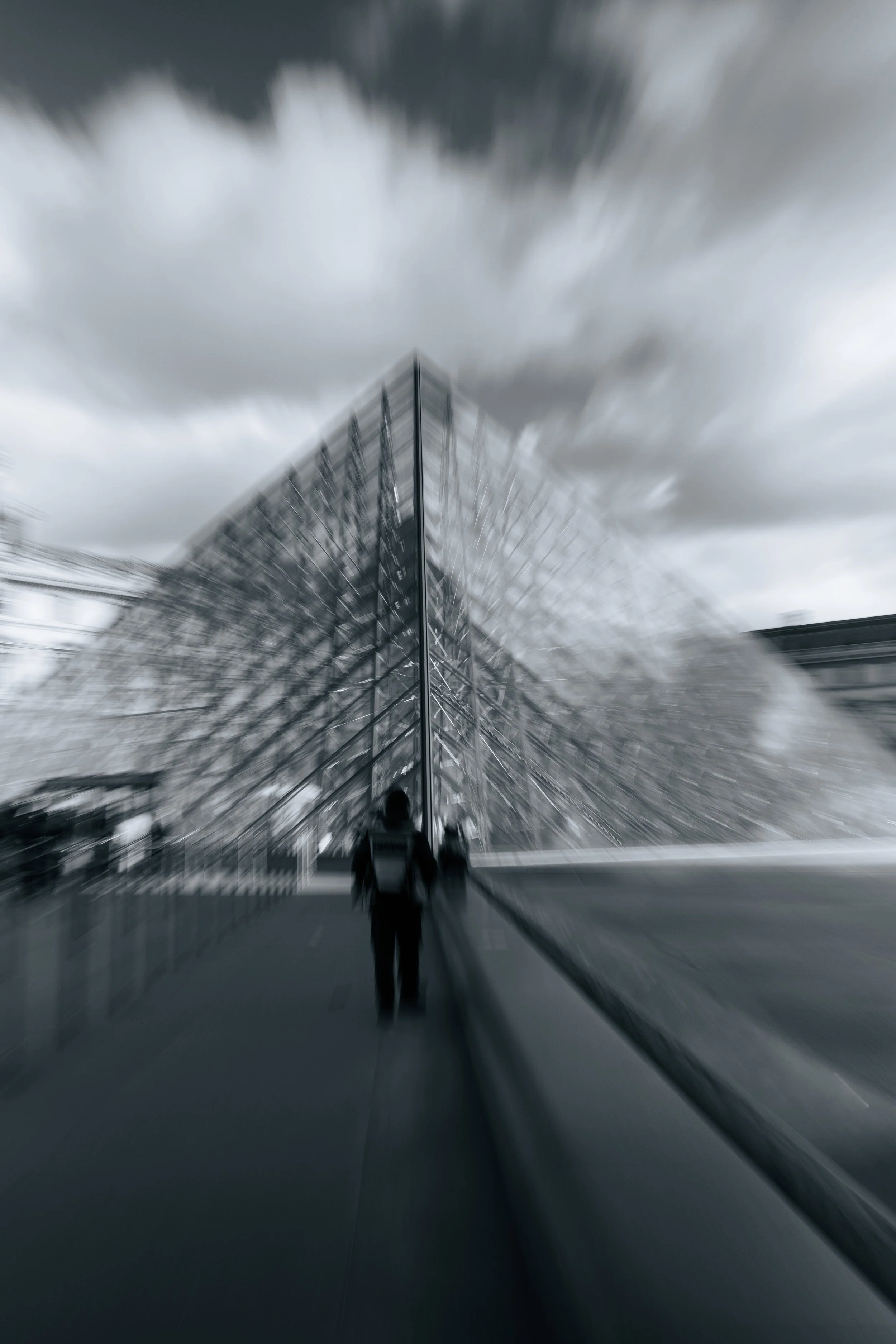 People walking near a modern glass building with a peaked roof under cloudy sky, with dramatic motion blur.