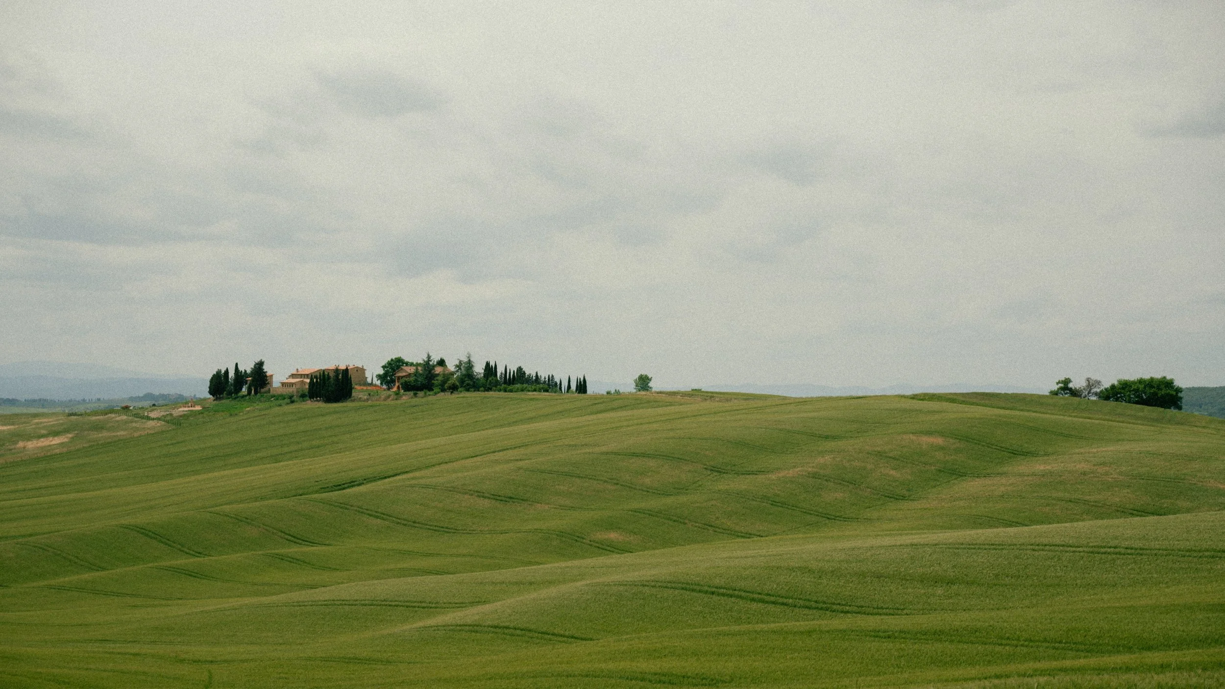Rolling green hills with a few houses and trees in the distance under a partly cloudy sky.