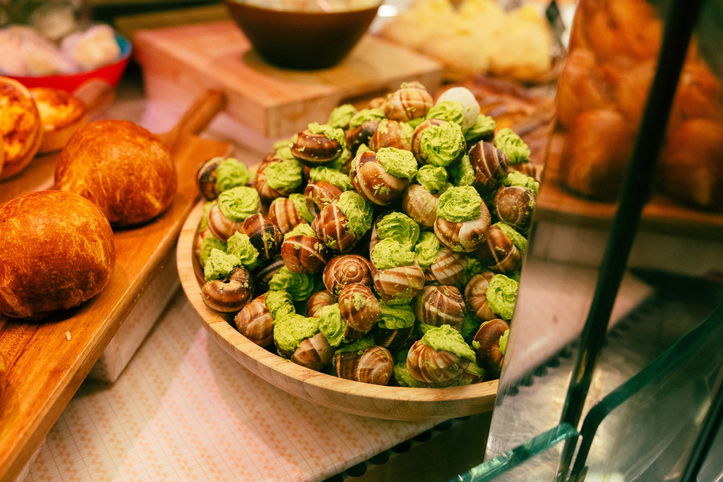 A wooden bowl filled with snails coated in green seasoning or sauce at a food market.