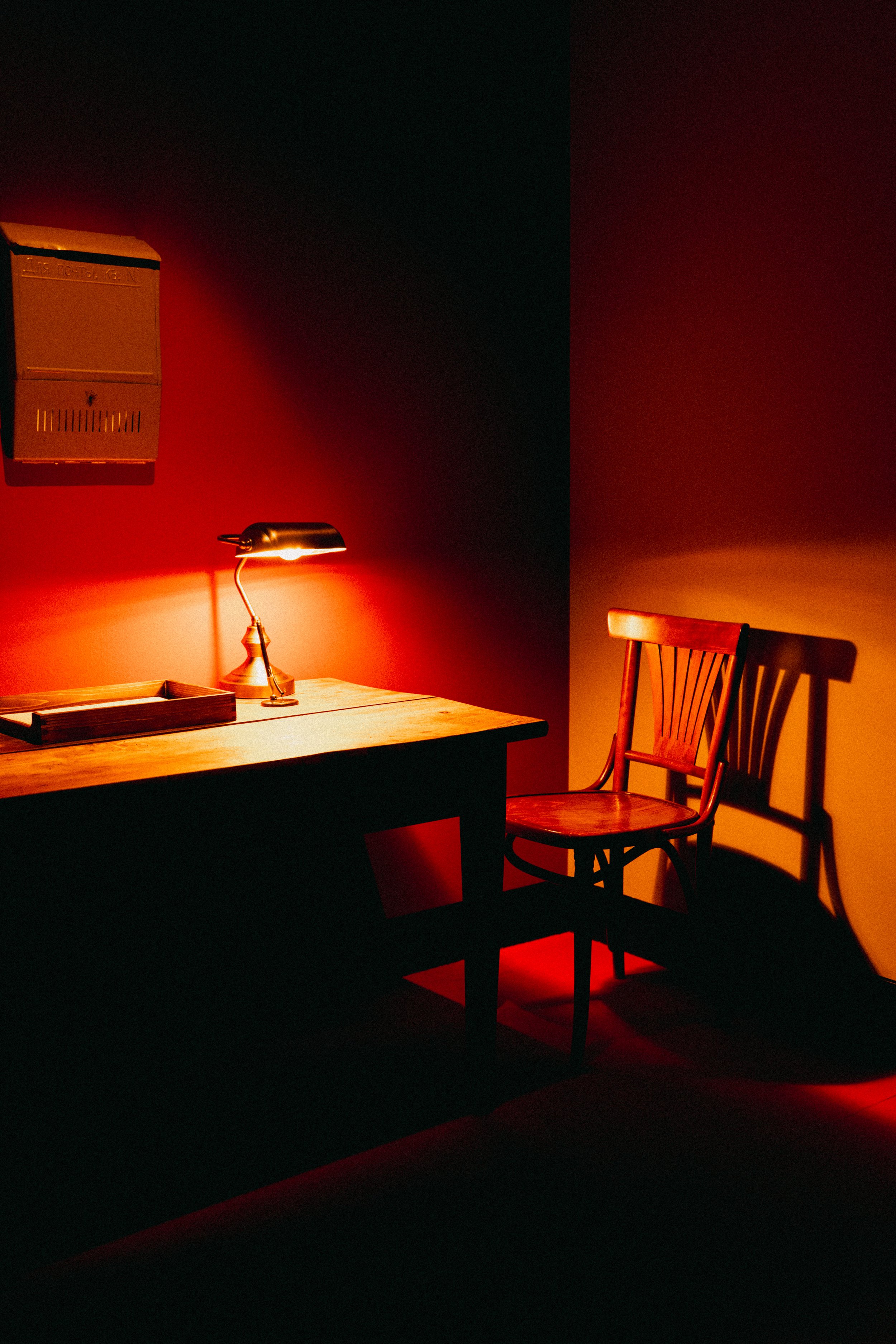 A dimly lit room with a wooden table, a desk lamp, a wooden chair, and a wall-mounted box, all illuminated with warm red and yellow lighting.
