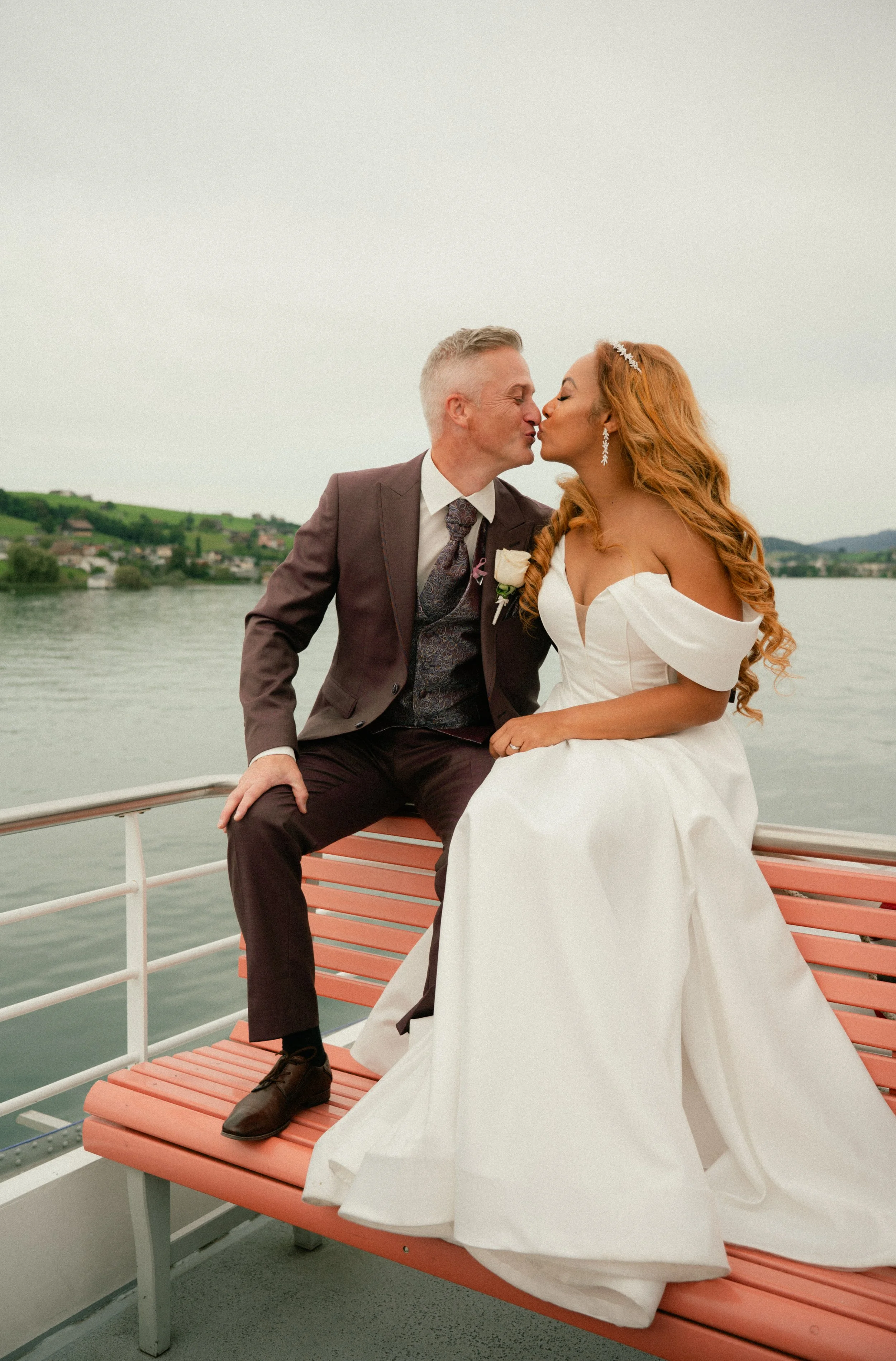 A newlywed couple sitting on a boat bench, about to kiss, with a river and green hills in the background.