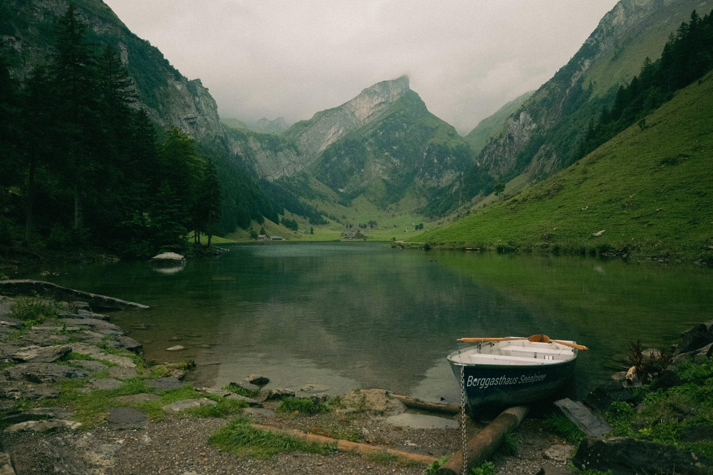 A peaceful mountain lake scene with green hills, tall mountains in the background, and a small boat named 'Berggasthaus Seelsee' on the rocky shore.