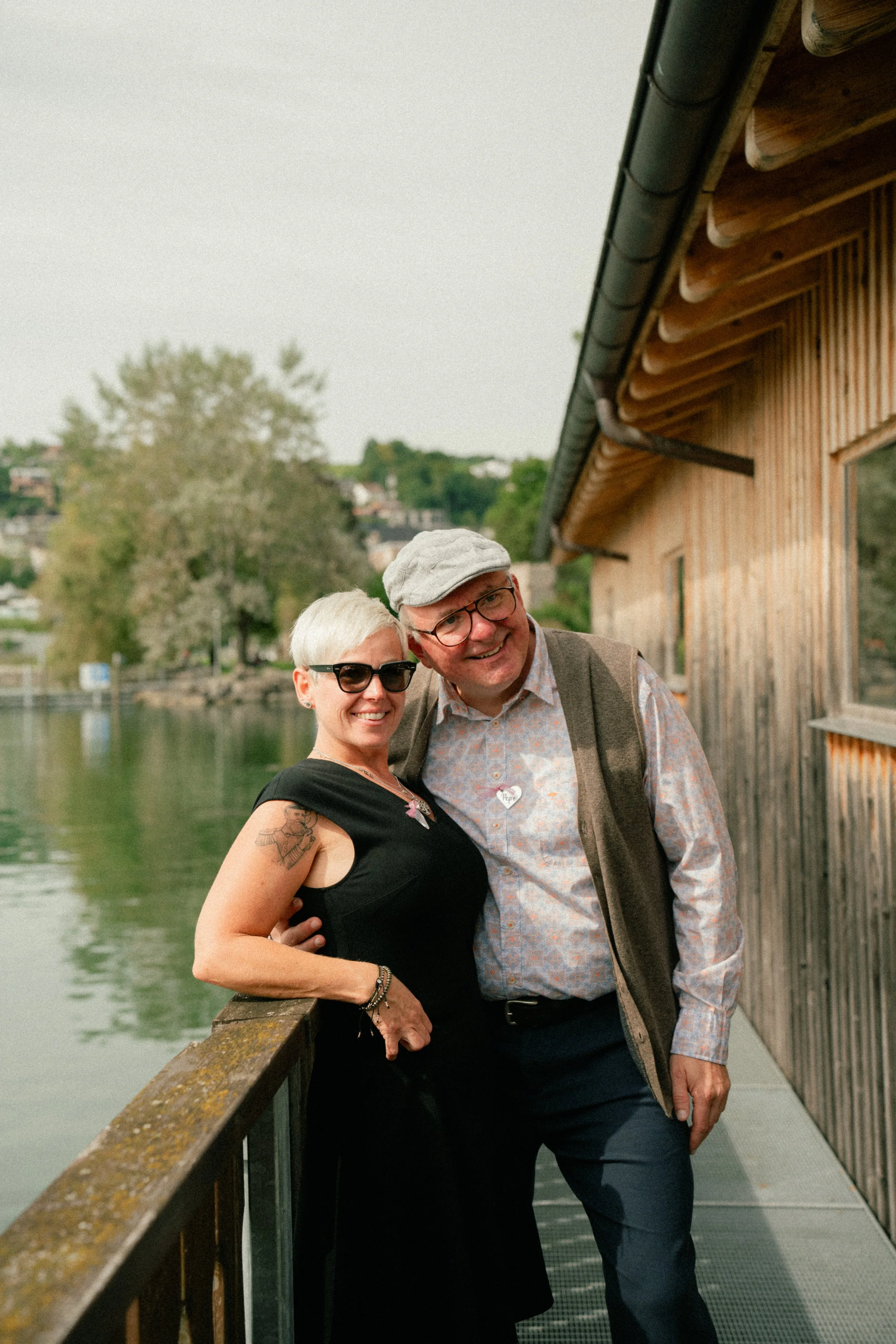 A smiling woman with short white hair and sunglasses standing next to a smiling man wearing a hat and glasses on a wooden dock by a lake.