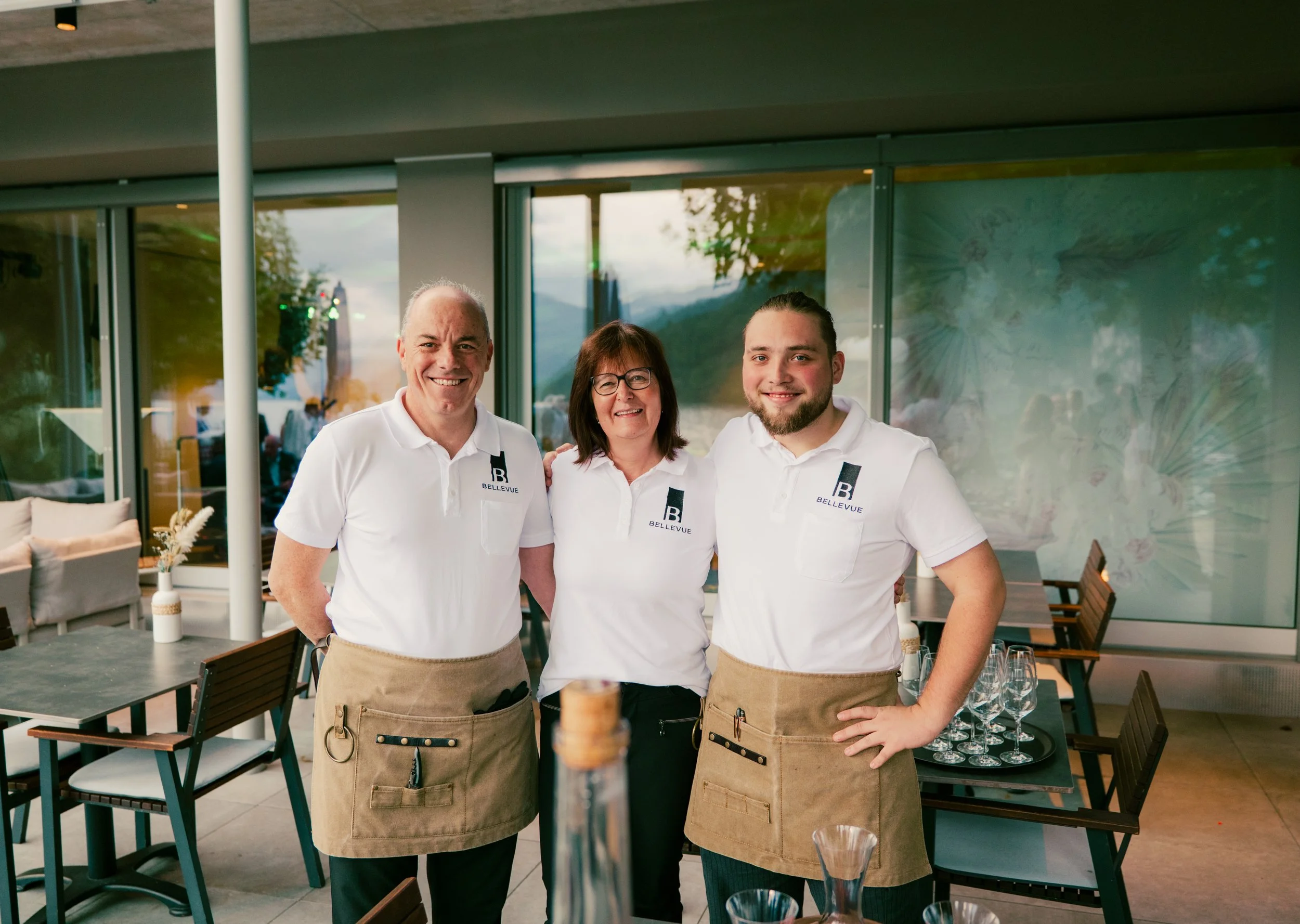 Three restaurant staff members wearing white polo shirts with 'B Bellevue' logo, standing indoors near a window, smiling at the camera.