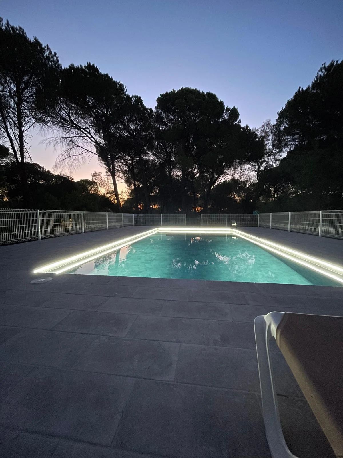 Piscina iluminada con luces LED en la tarde o noche, rodeada de una cerca de metal, con árboles en el fondo y un cielo despejado.