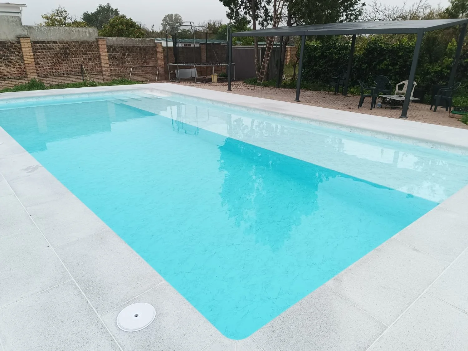 Piscina rectangular con agua azul, rodeada de baldosas grises, en un área al aire libre con vista a un muro de ladrillos, árboles y mobiliario de jardín, incluyendo sillas y una estructura con cobertura