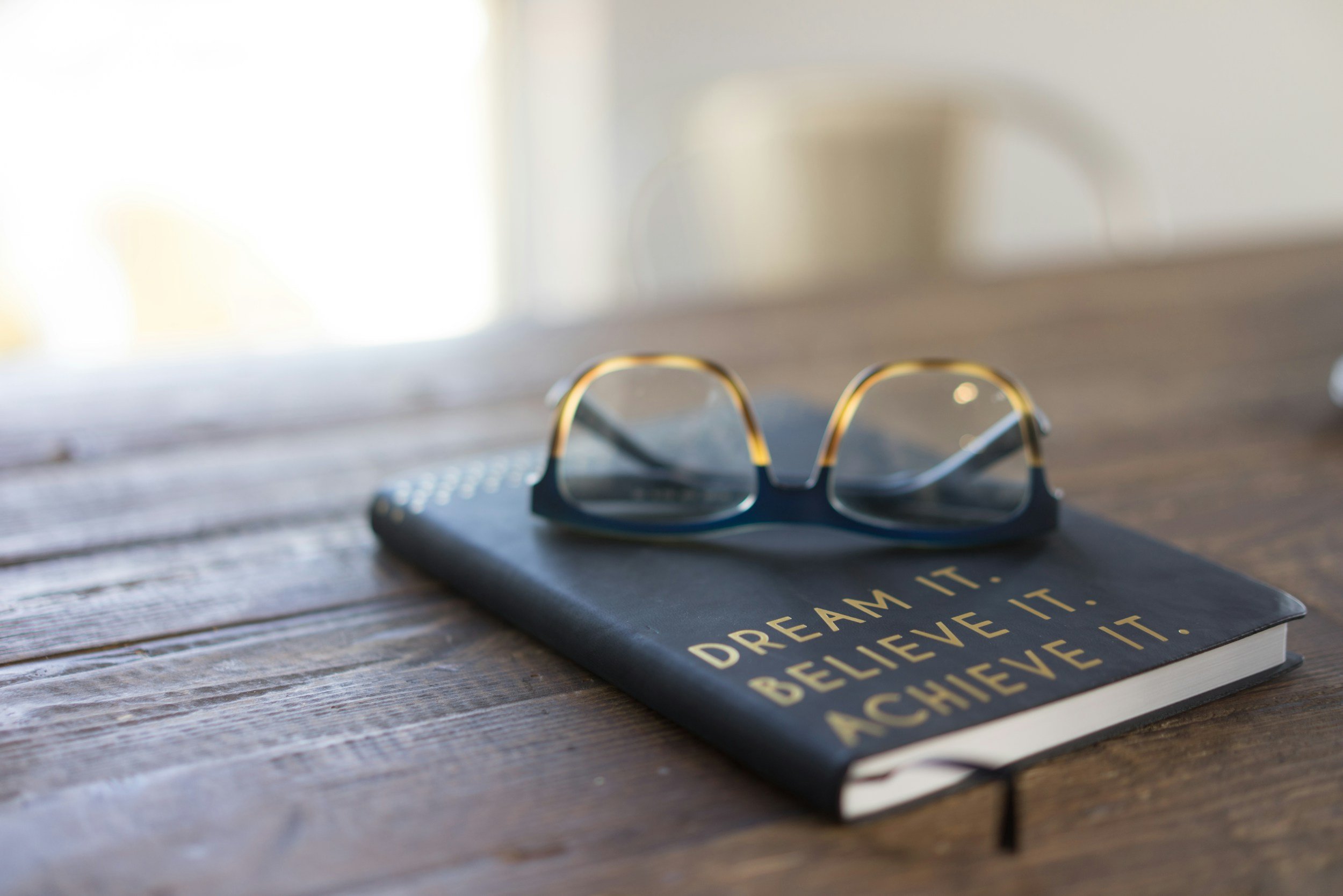 A pair of eyeglasses resting on a closed notebook with the motivational quote, 'Dream it. Believe it. Achieve it.' printed on the cover, placed on a wooden table.