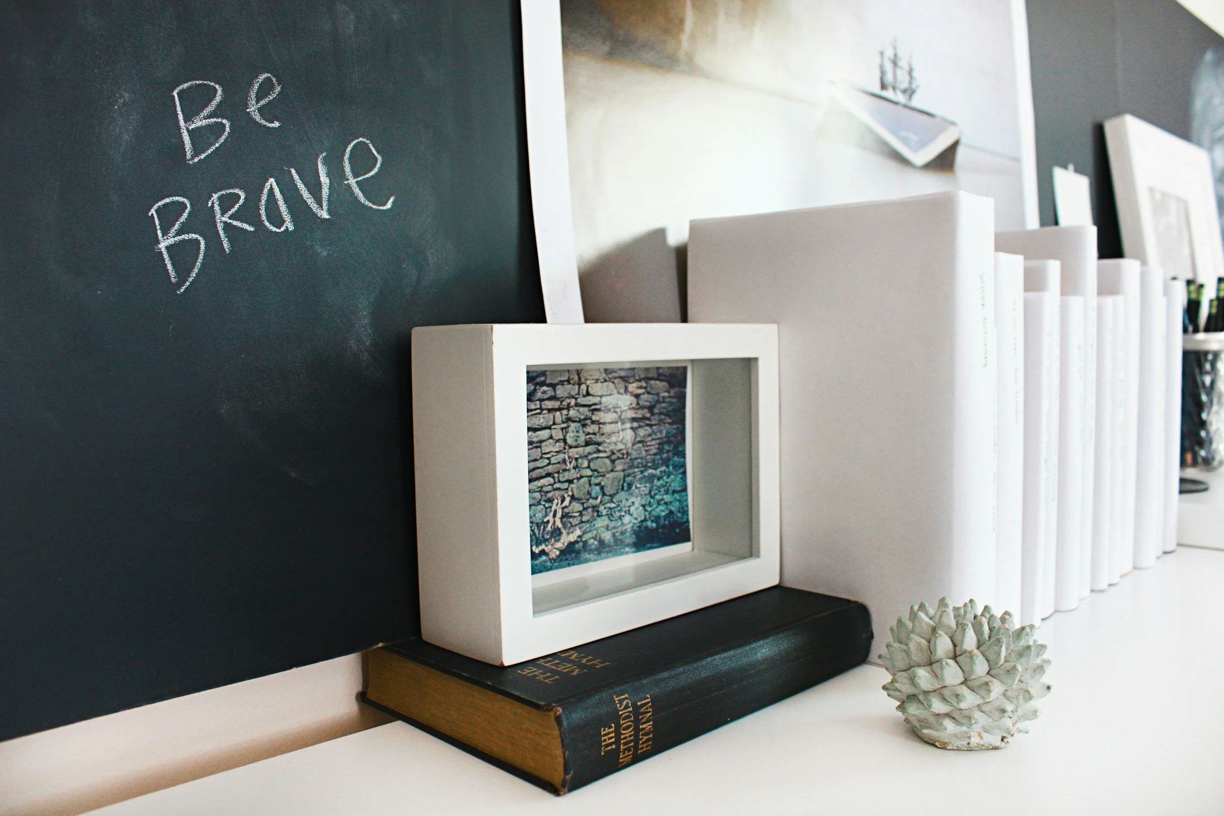 A black chalkboard with the words "Be Brave" written in white chalk, a white picture frame with a photo of a stone wall and water, a stack of white folders or books, a black book titled "The Ethicist" under the picture frame, and a small decorative white pine cone on a white surface.