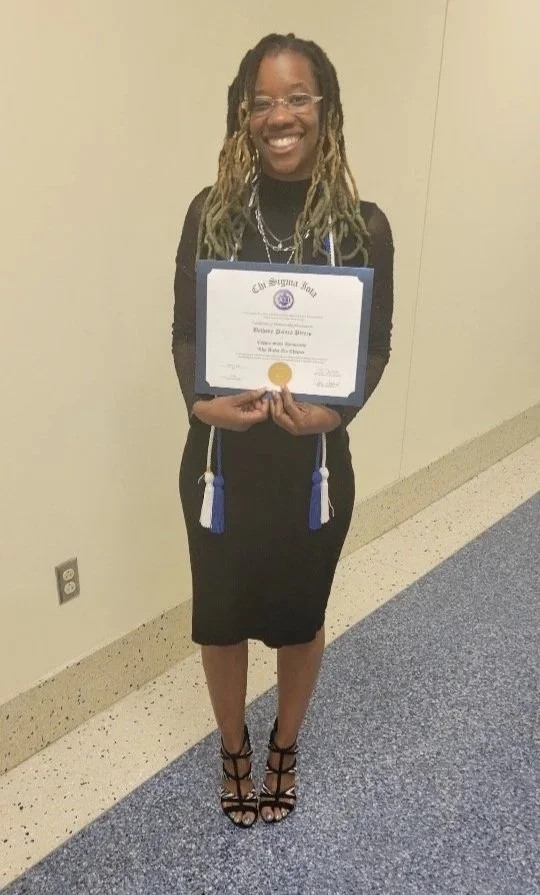 A woman with glasses and long dreadlocks smiling while holding a graduation diploma, wearing a black dress and high heels, standing against a beige wall.