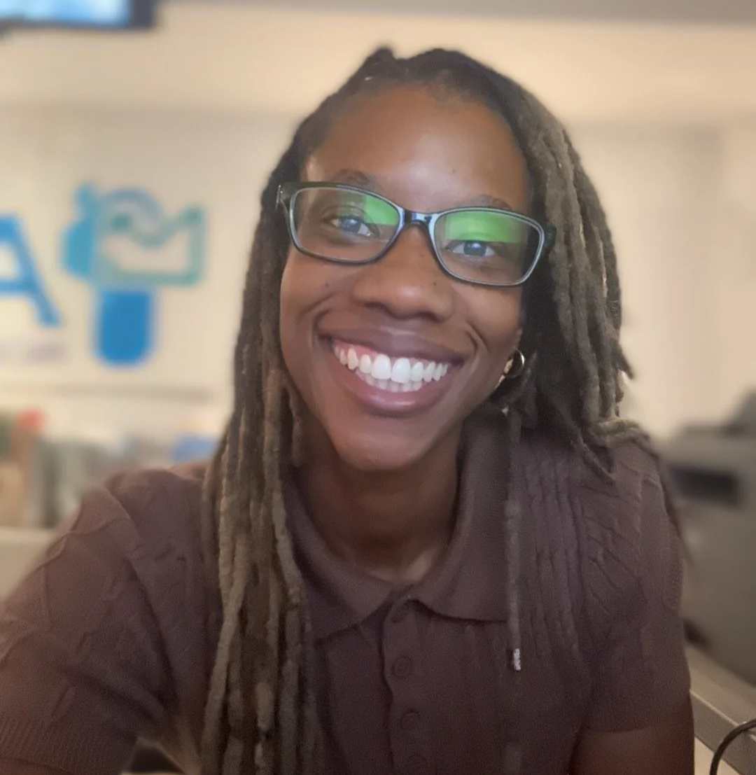 A smiling woman with glasses and dreadlocks, wearing a brown polo shirt, sitting indoors with a blurred background that includes a blue logo on the wall.