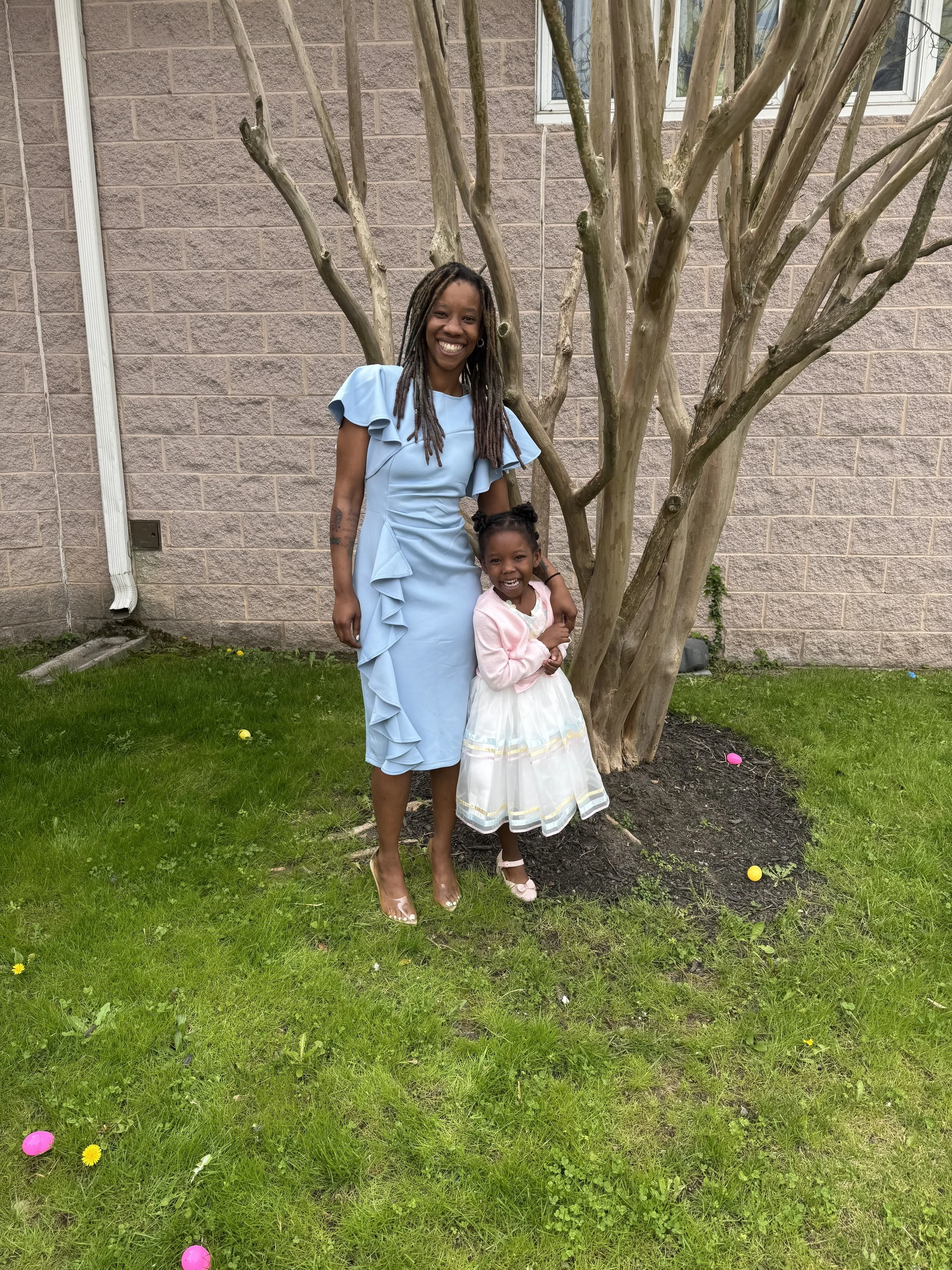A woman and a young girl smiling and standing together outdoors near a tree, with a brick building in the background, surrounded by green grass and colorful plastic eggs on the ground.