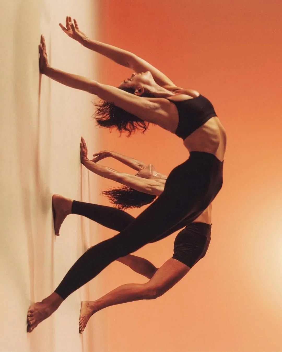 Two women practicing yoga on a beige floor with an orange gradient background, performing a pose with their hands and feet on the ground, stretching their bodies.