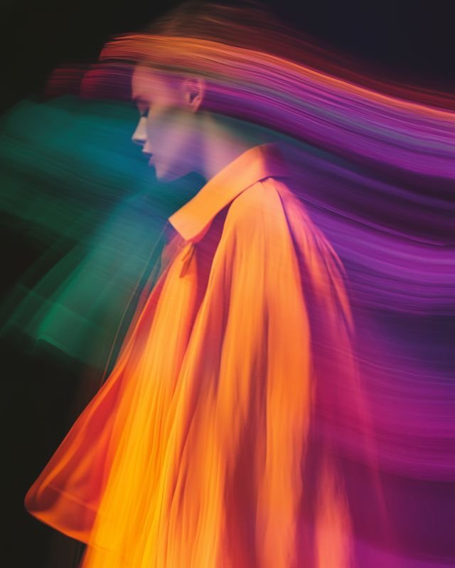 A woman in a yellow-orange coat with dynamic rainbow-colored light streaks surrounding her