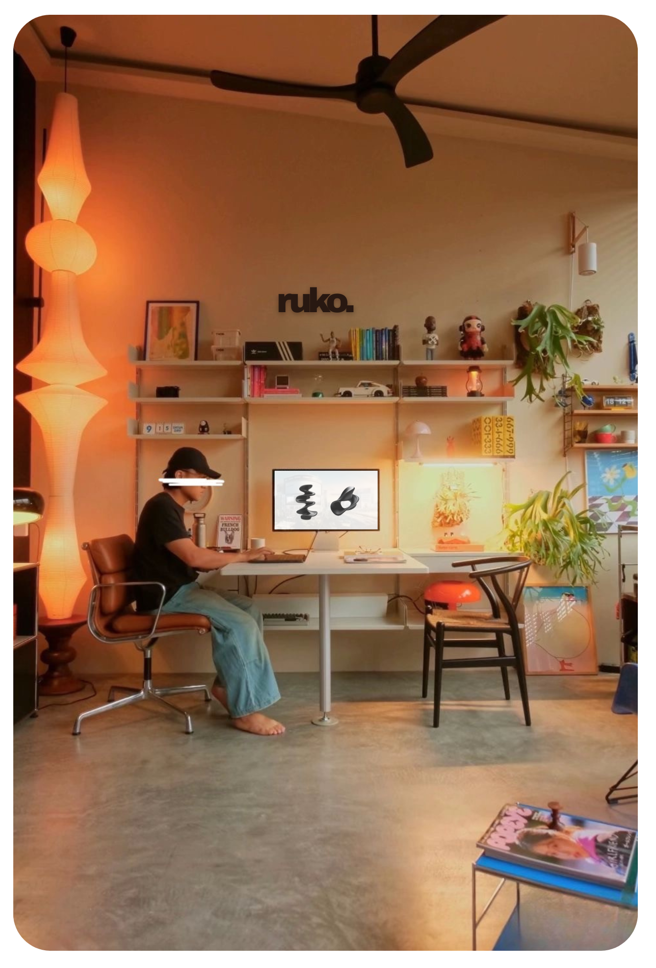 Living room with a person sitting at a desk, working on a laptop. The room is decorated with shelves, artwork, plants, and a television displaying abstract black and white shapes. Warm lighting and modern decor create a cozy atmosphere.