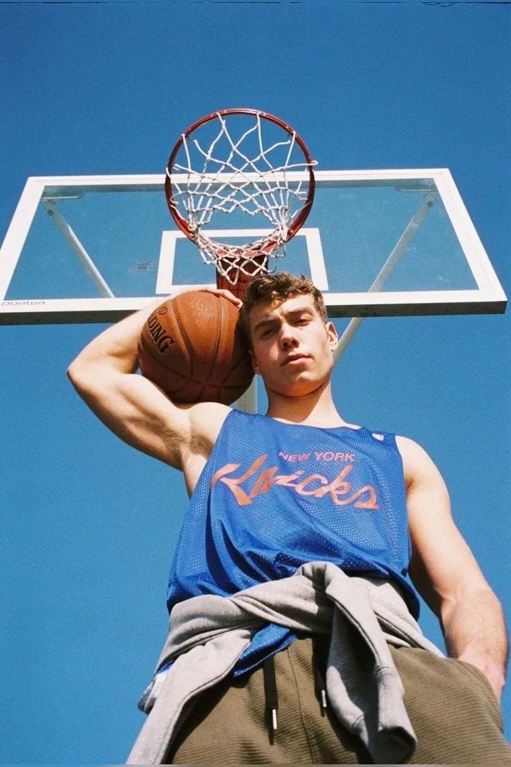 Young man in a blue basketball jersey holding a basketball, standing outside under a basketball hoop against a clear blue sky.