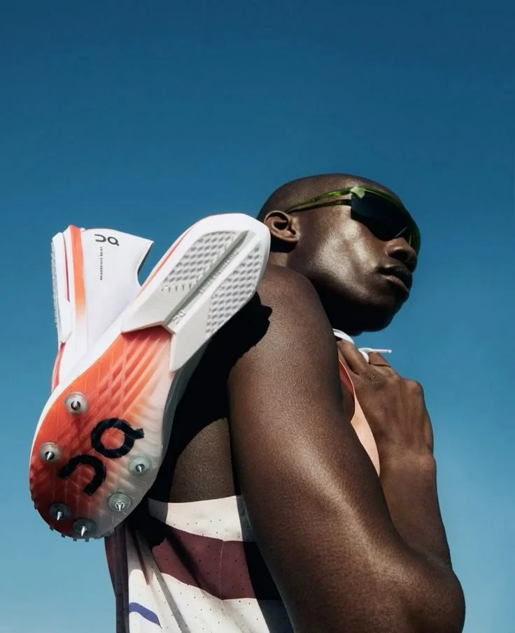 A male athlete wearing sunglasses and a sports tank top, carrying a white and orange running shoe on his shoulder against a clear blue sky.