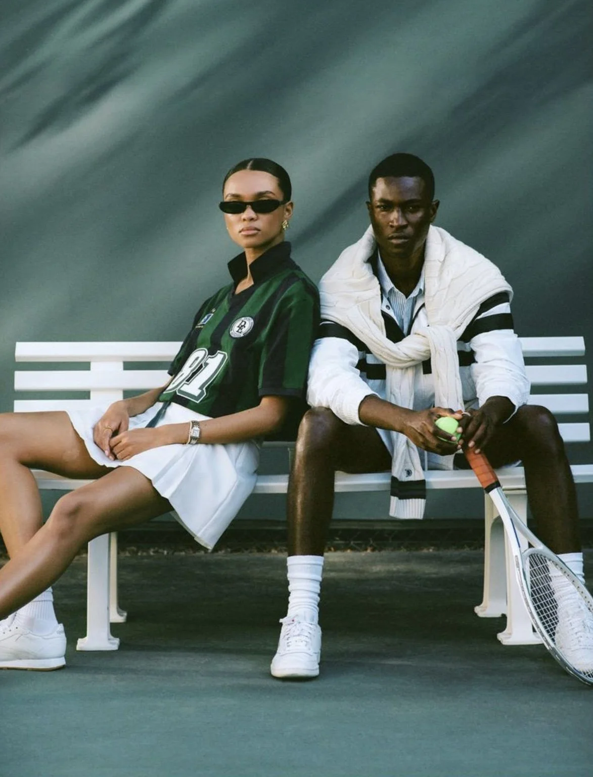 A man and woman sitting on a white bench on a tennis court. The woman is wearing a green and black sports jersey, white shorts, and sunglasses. The man is holding a tennis ball and racket, wearing a striped shirt, white jacket tied around his shoulders, and white sneakers.