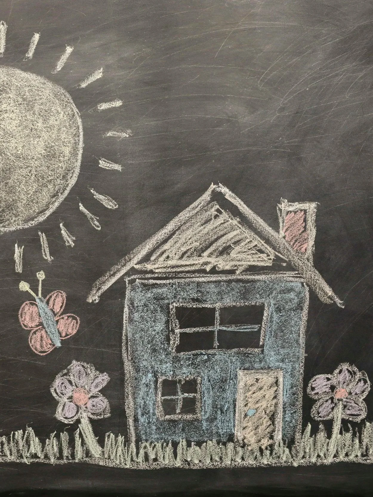 A chalk drawing of a house with a triangular roof, chimney, four windows, and a door, surrounded by flowers and a butterfly on a dark background.